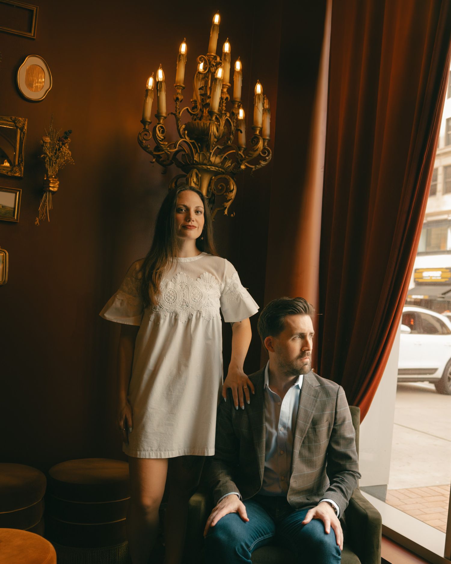 A woman in white dress and man in gray suit pose elegantly beneath a golden chandelier in a warmly lit room.