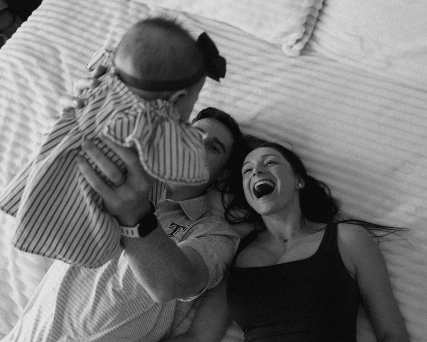Black and white photo of a laughing woman and man holding a baby on a bed, capturing a joyful family moment.