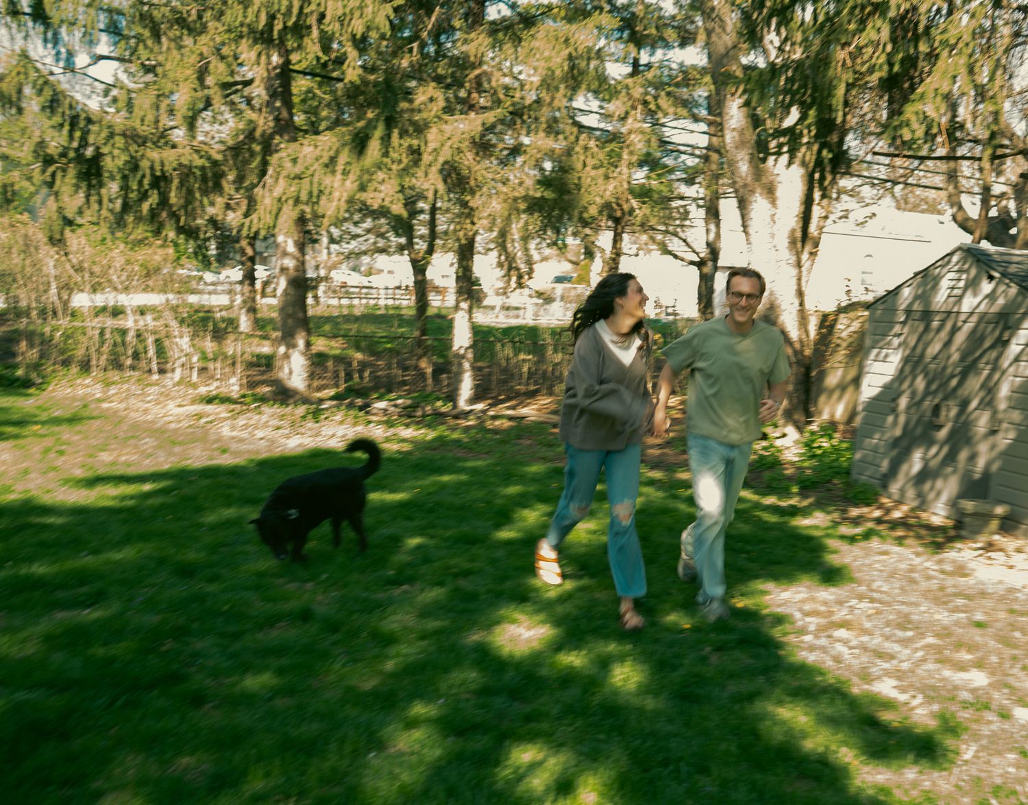 Couple walking together on green lawn with black dog in sunny backyard surrounded by tall trees.