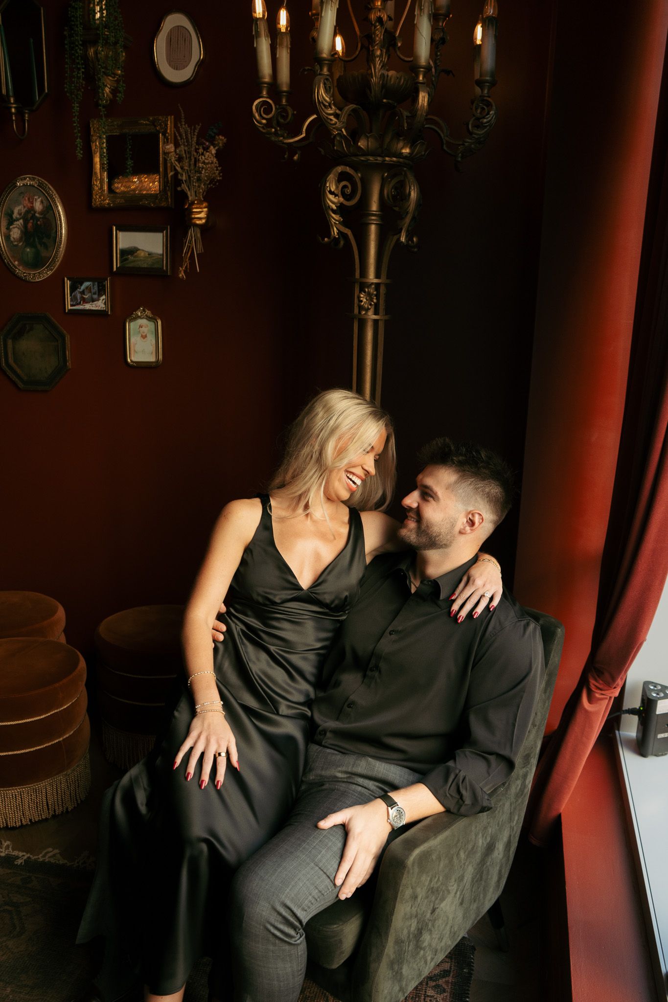 Elegant couple in black attire sharing a romantic moment in a dimly lit, vintage-styled room with chandelier.