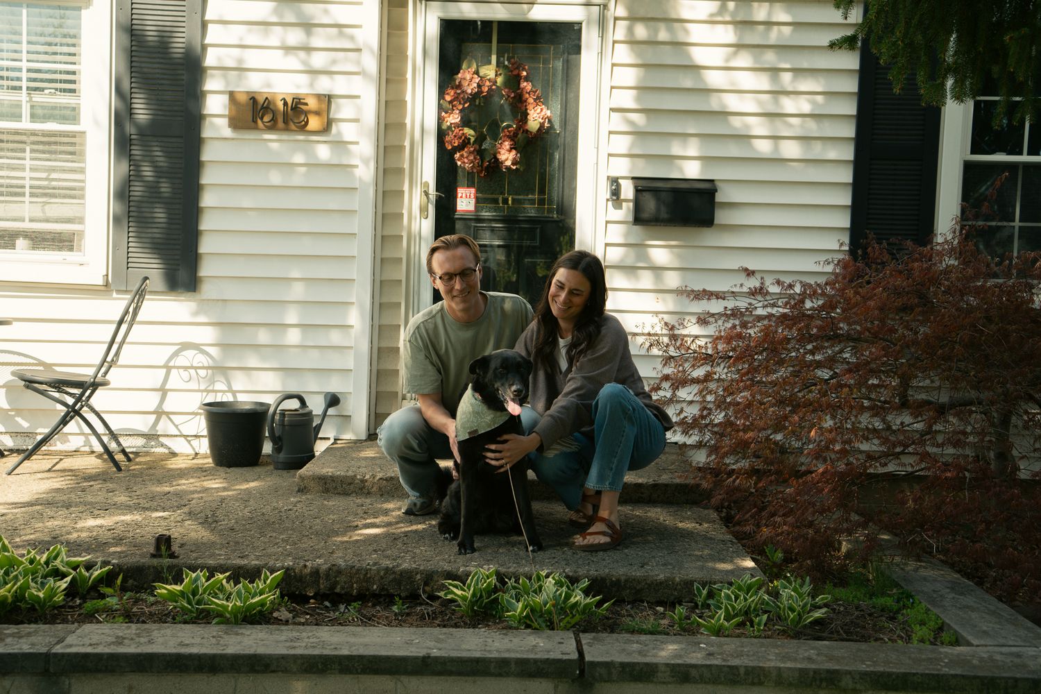 Couple sitting on front porch steps with black and white dog outside white house with wreath on door.