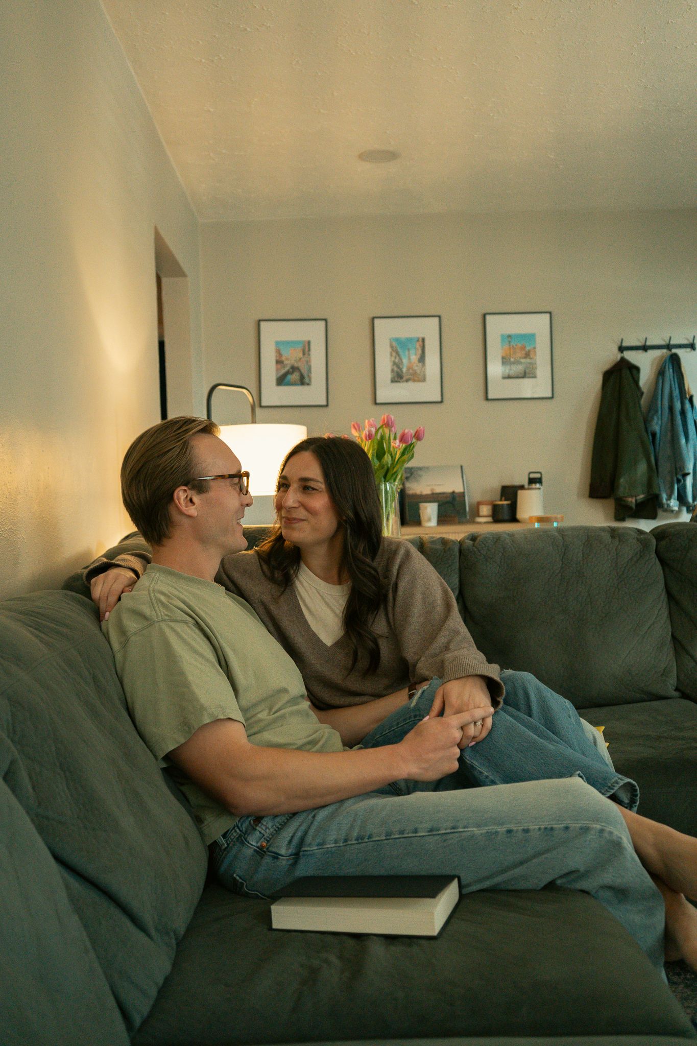 Couple sitting closely on a gray couch, smiling at each other in a cozy living room with framed art and tulips.