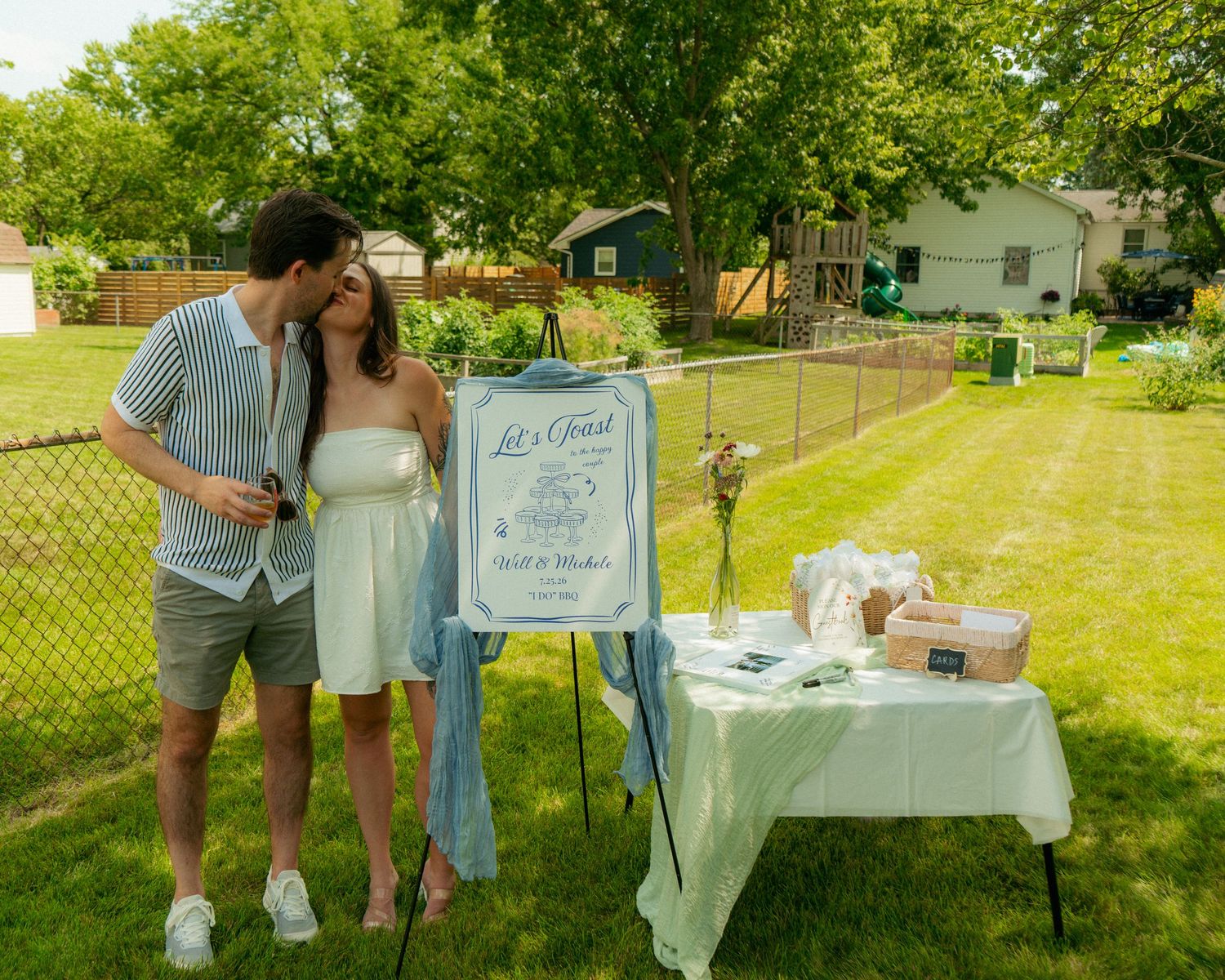 Couple posing by a wedding guest book table at an outdoor backyard celebration on a sunny day.