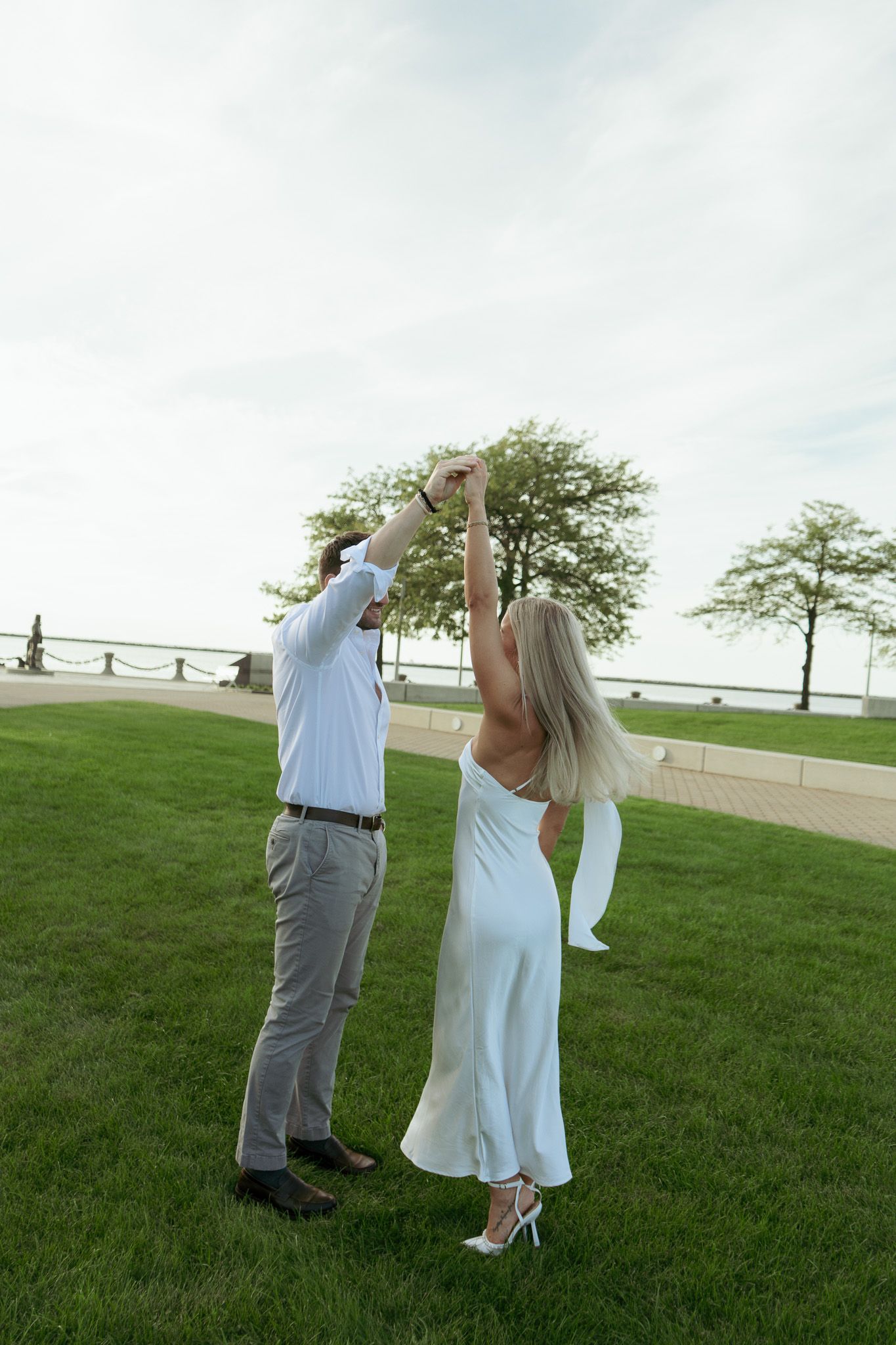 A couple dances outdoors on green grass, woman in white dress with arms raised, trees and water visible in background.