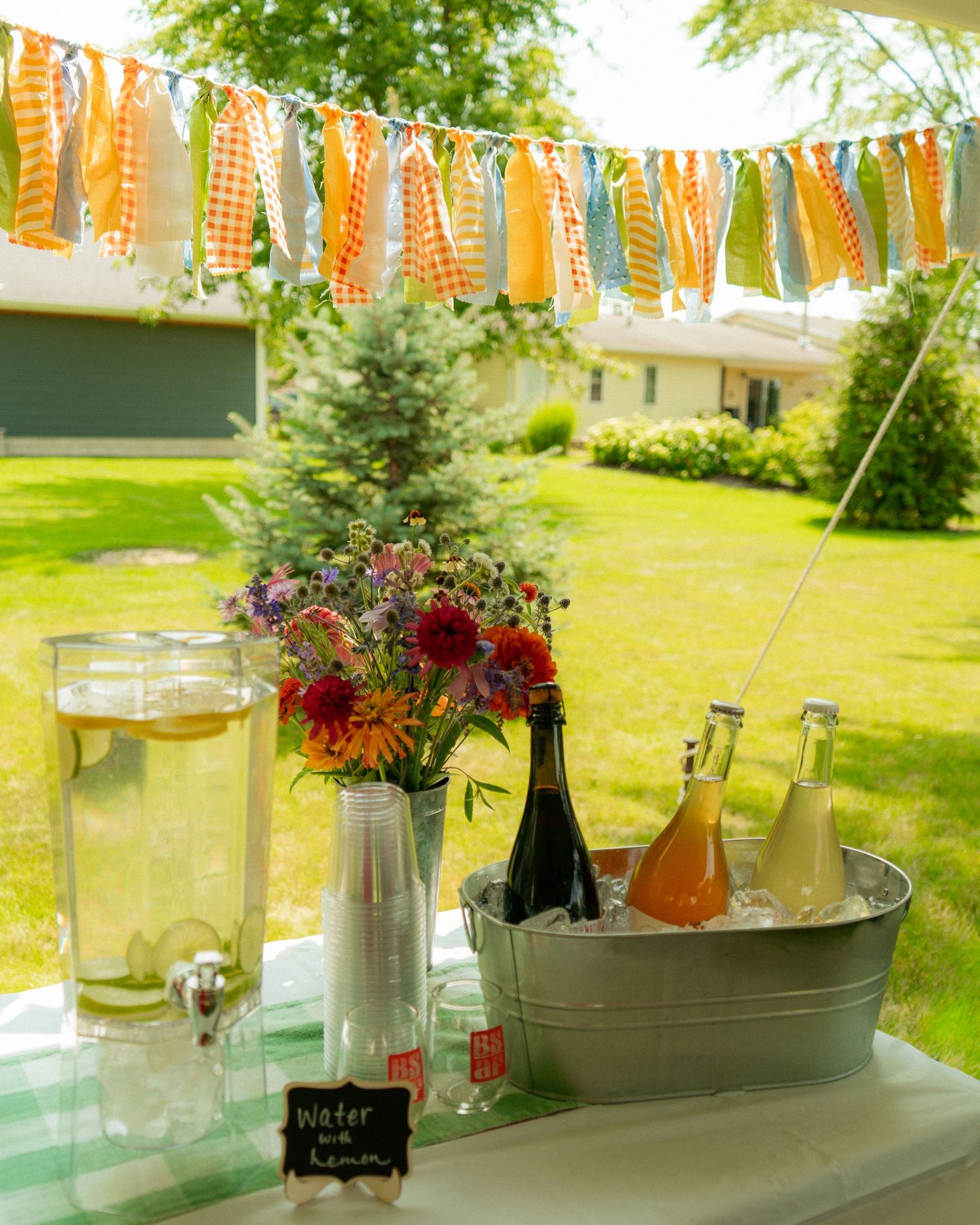 Outdoor party drink station with lemonade, bottled beverages in ice bucket, fresh flowers, and colorful bunting banner.