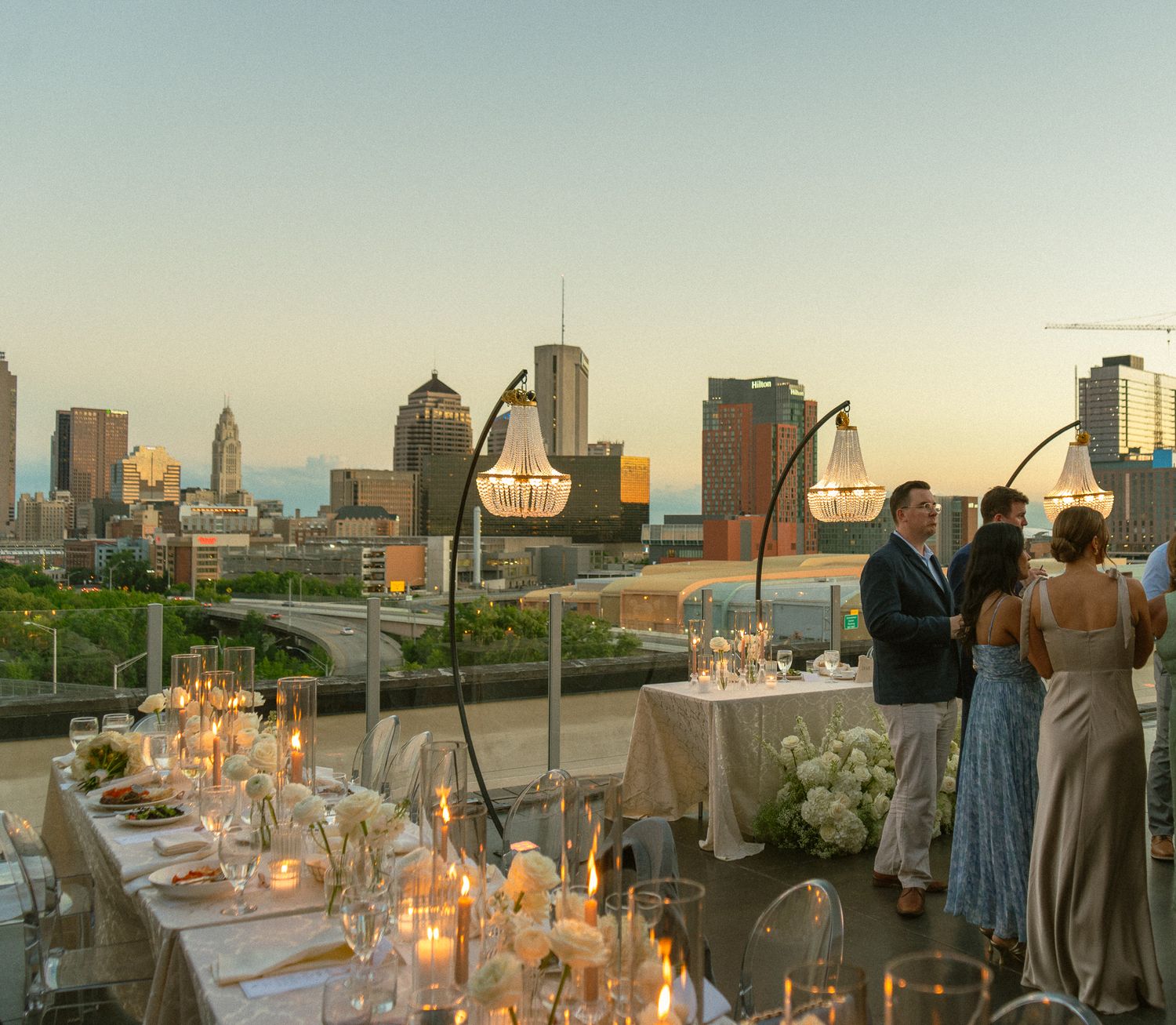 Rooftop wedding reception with candlelit tables and city skyline at sunset, guests mingling near elegant chandeliers.