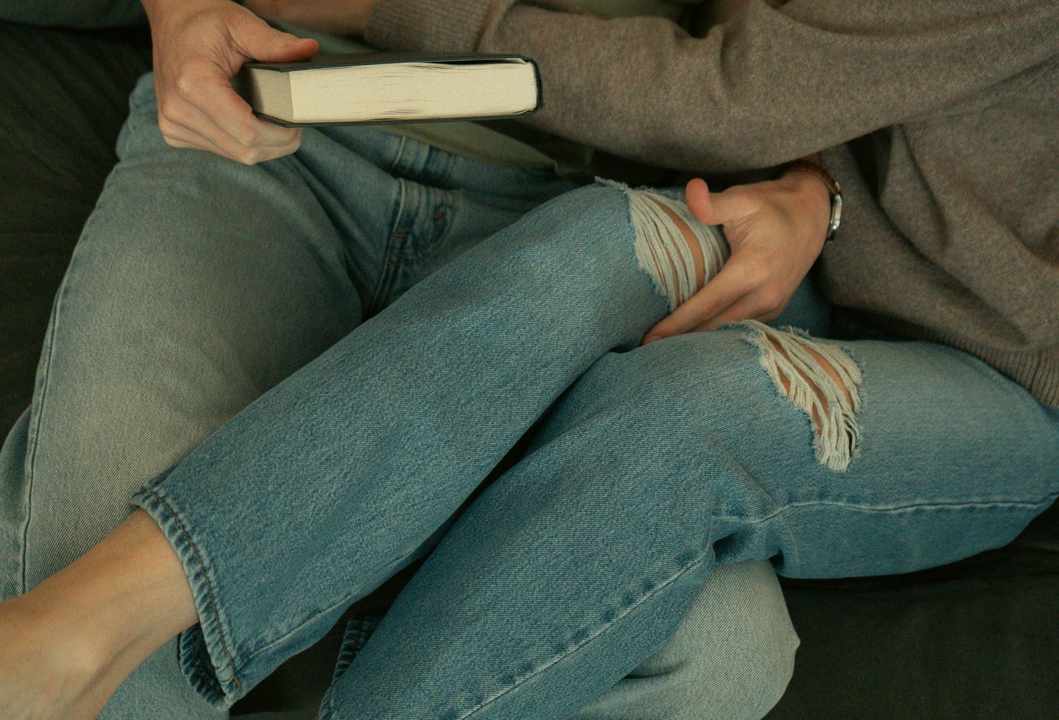 Two people sitting together on a couch, one holding a book, both wearing ripped blue jeans.