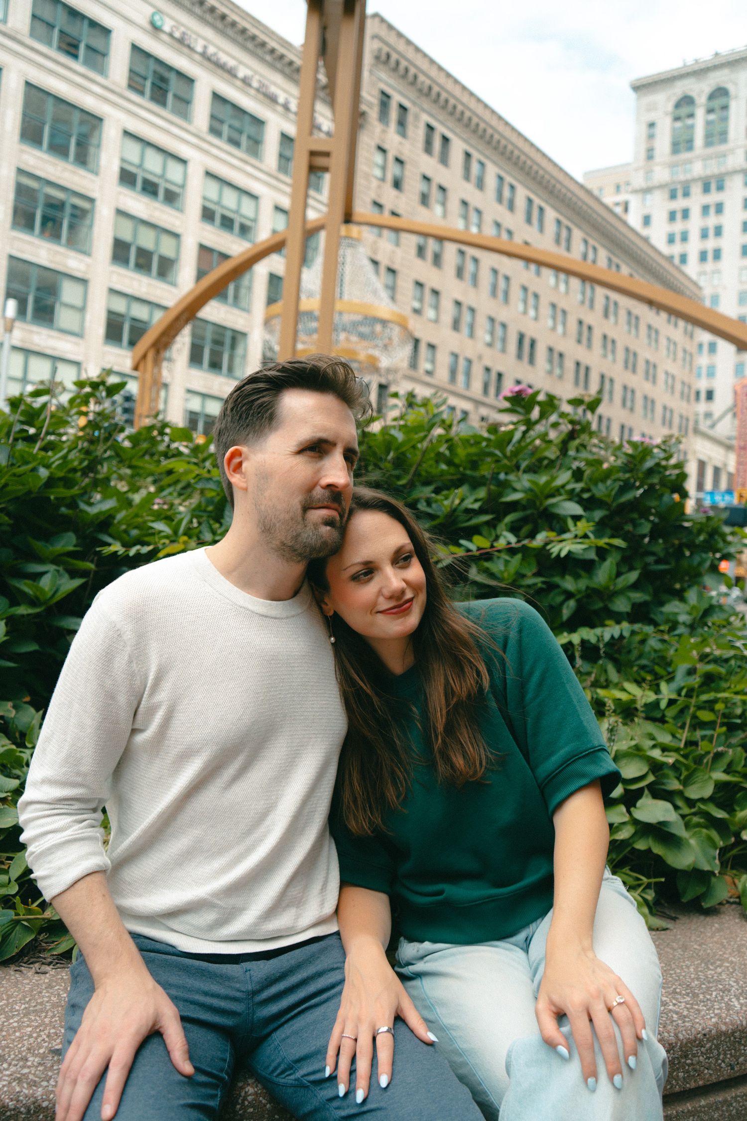Couple sitting together outdoors in an urban setting with lush greenery and city buildings behind them.