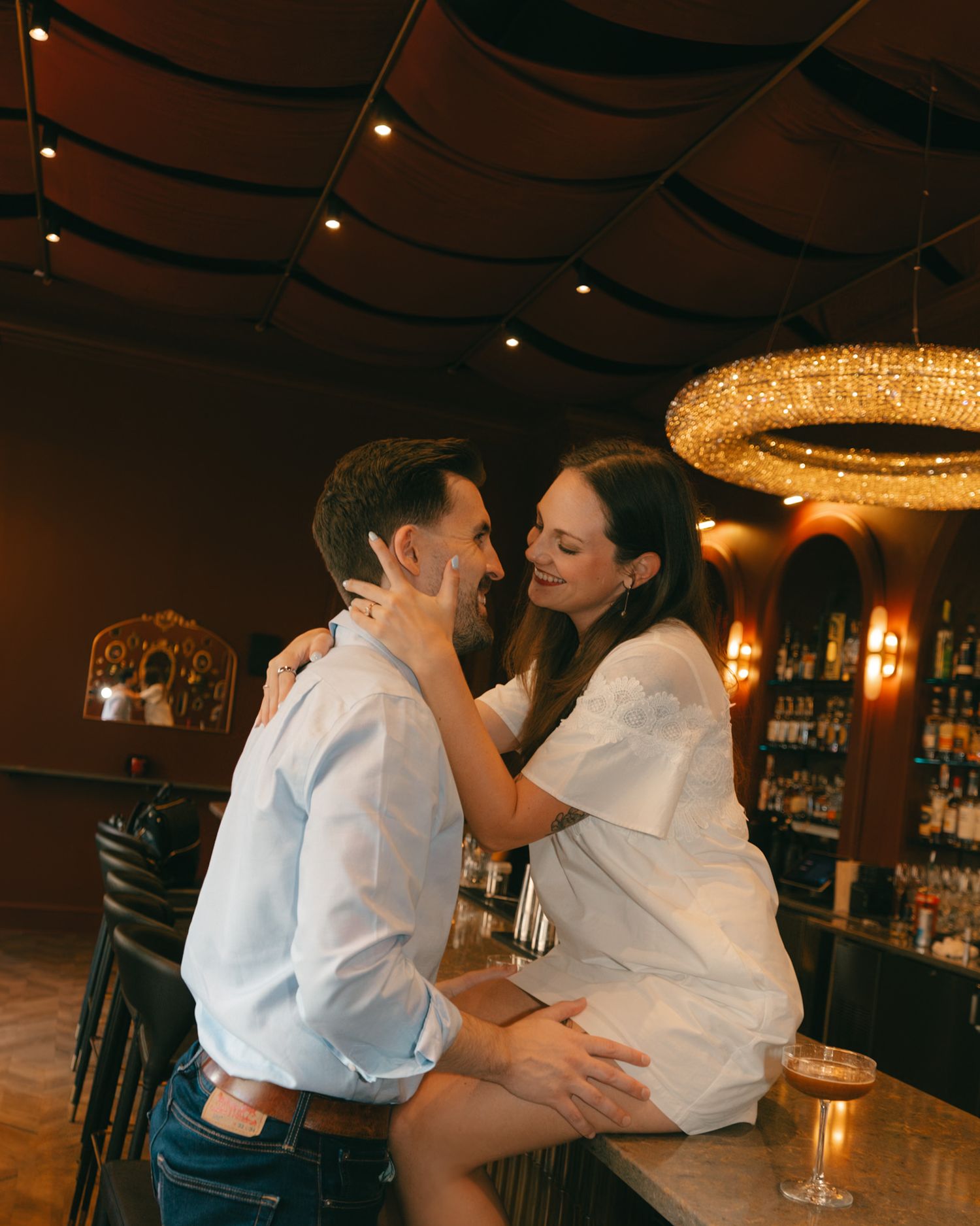 Couple sharing a romantic moment at a dimly lit bar with a chandelier overhead, both dressed in white.