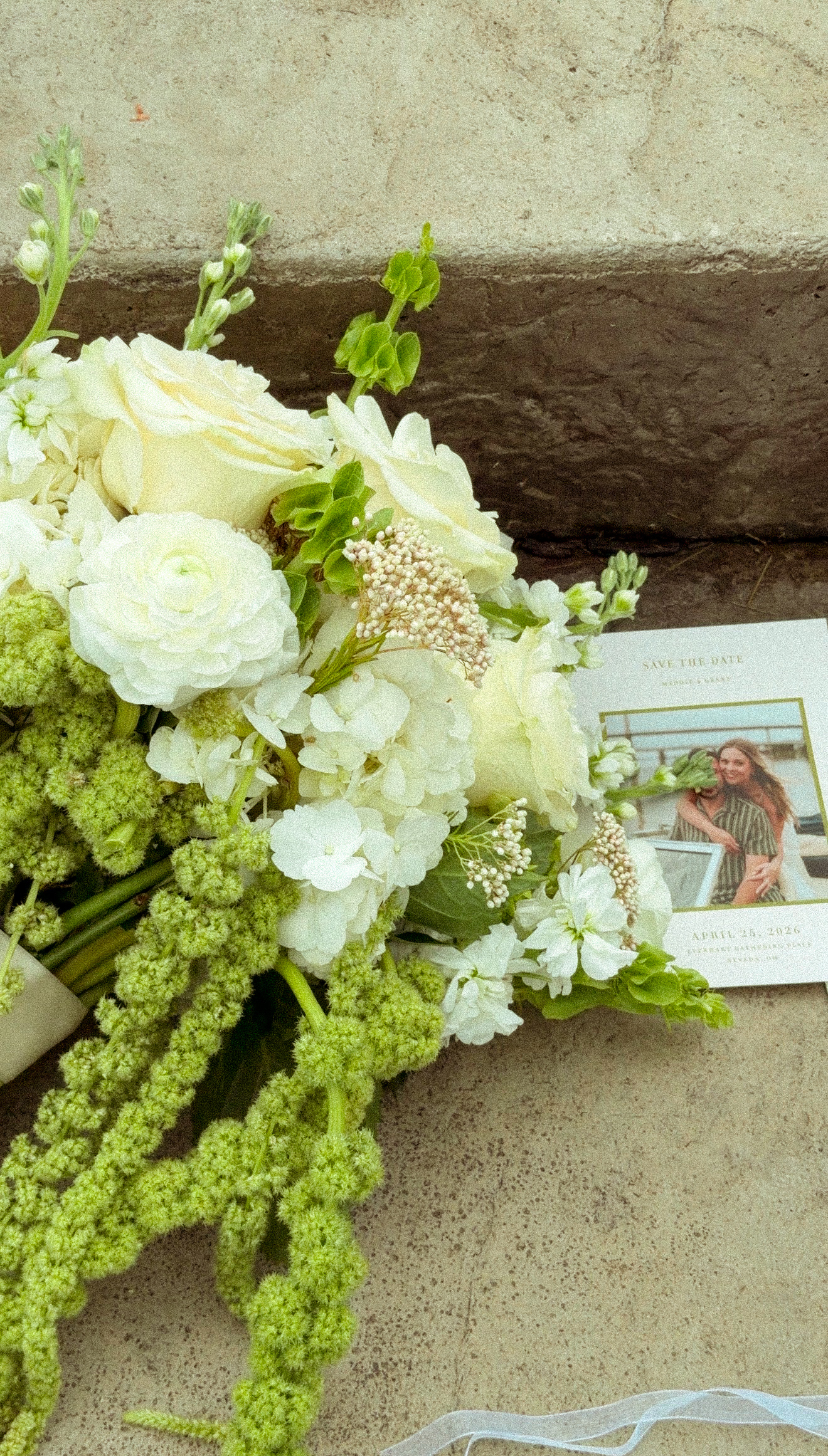 White roses and green amaranthus bridal bouquet resting on stone steps beside a wedding program card.