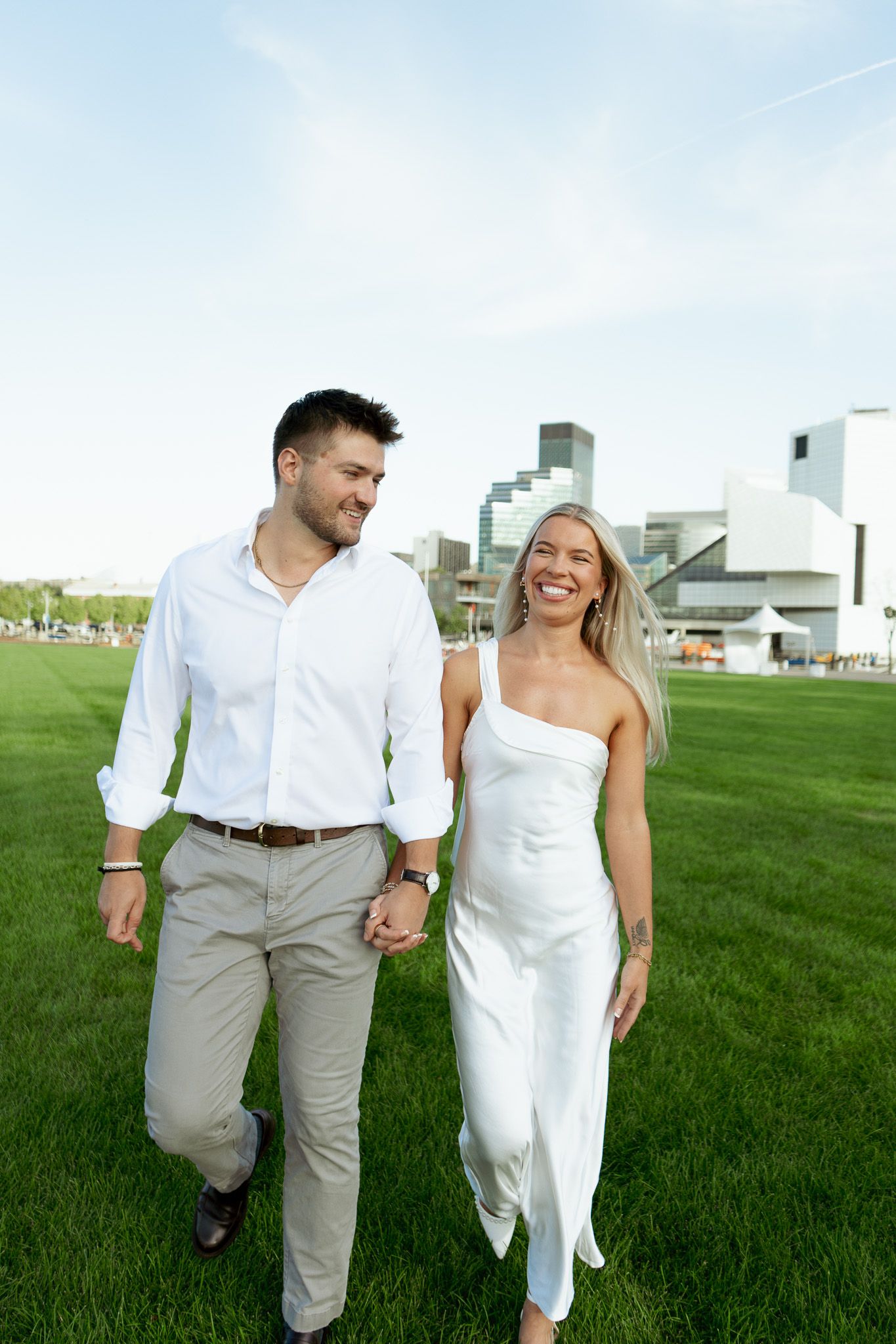 A smiling couple walks hand-in-hand on green grass, wearing white outfits, with a modern building skyline behind them.