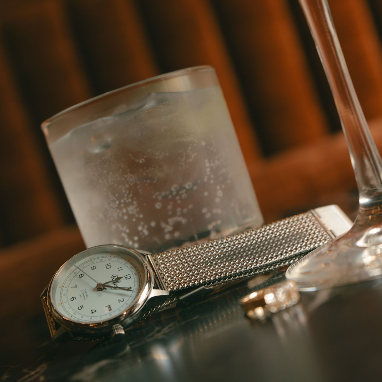 A silver mesh wristwatch rests beside a glass of water with ice on a dark reflective surface.