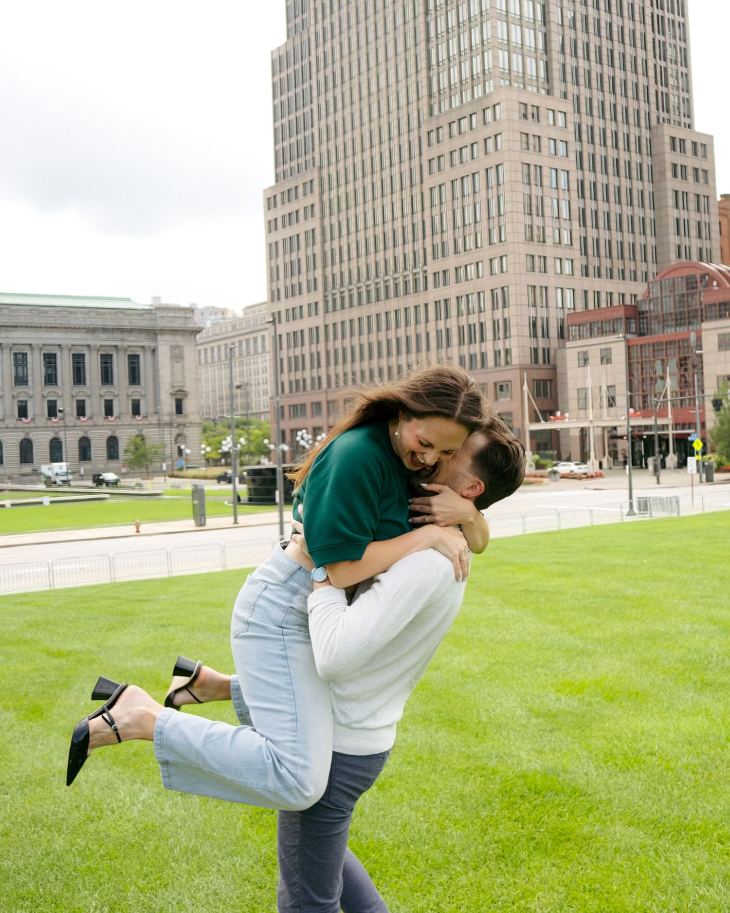 Romantic couple embracing as man lifts woman off ground in urban park with tall buildings in background.