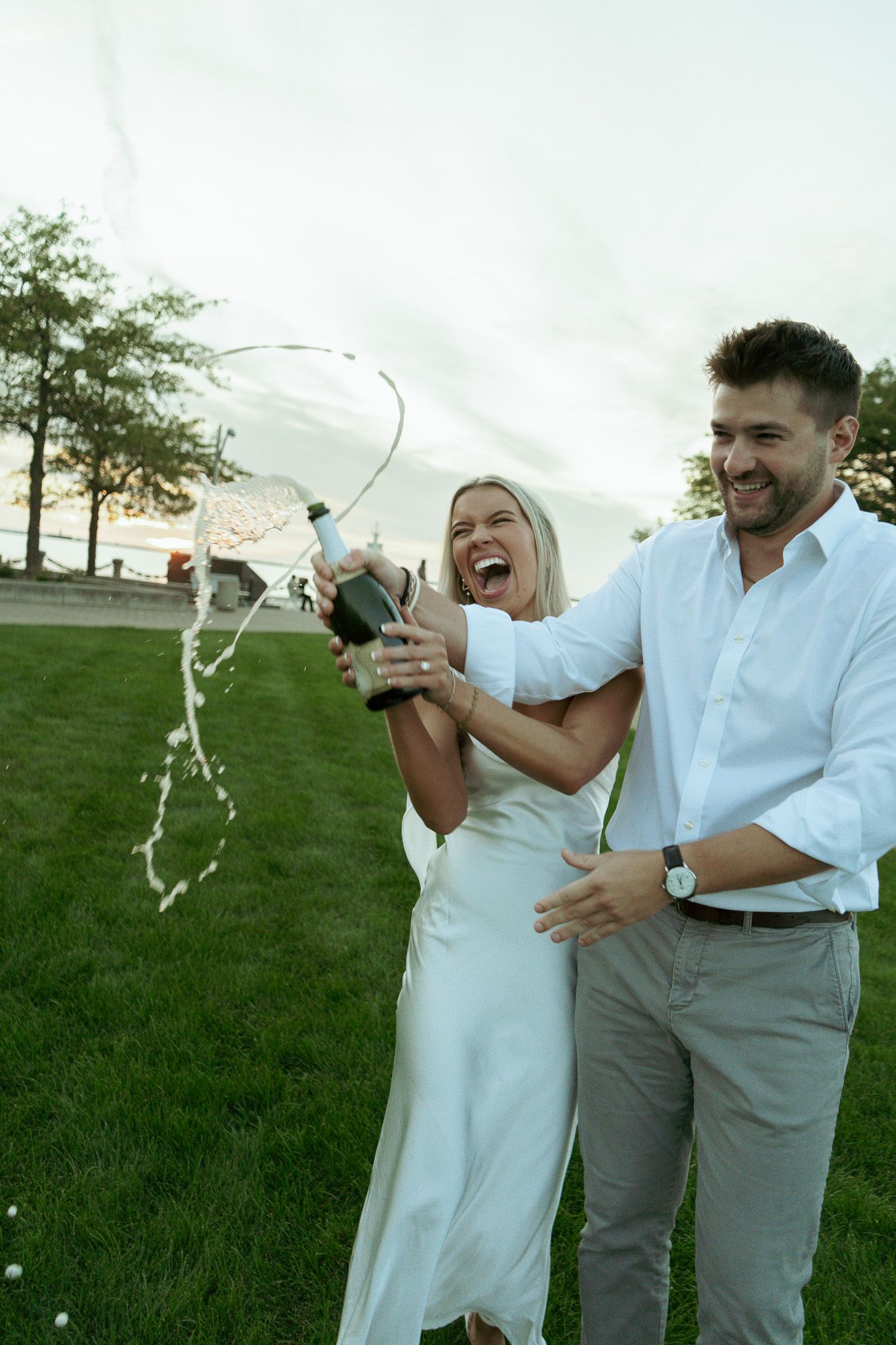 A joyful couple celebrates outdoors, popping champagne on a green lawn, the woman in white dress laughing as bubbles spray.