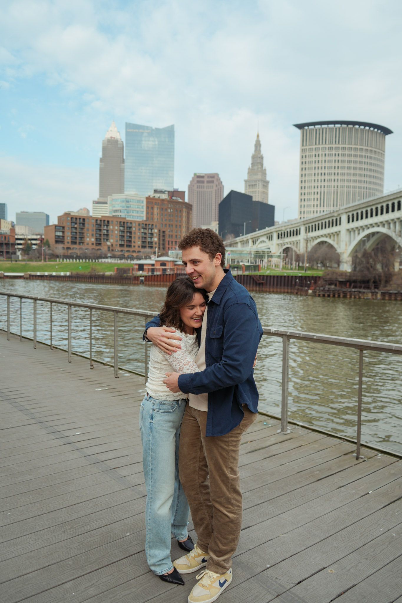 A couple shares a romantic moment on a wooden boardwalk along Cleveland's waterfront with the city skyline in view.
