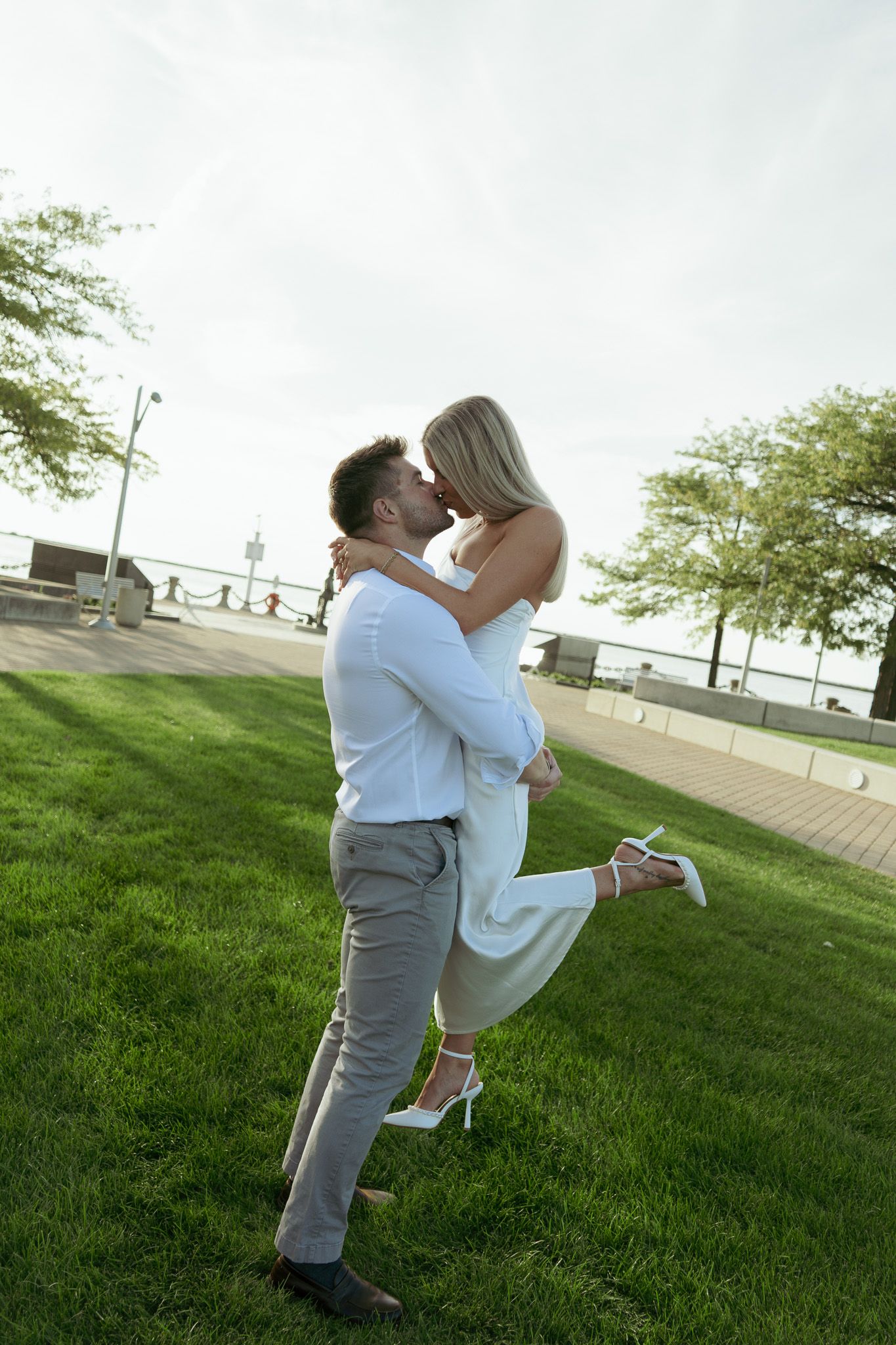 Couple sharing a romantic kiss on green grass near waterfront, woman in white dress lifted by man in white shirt.
