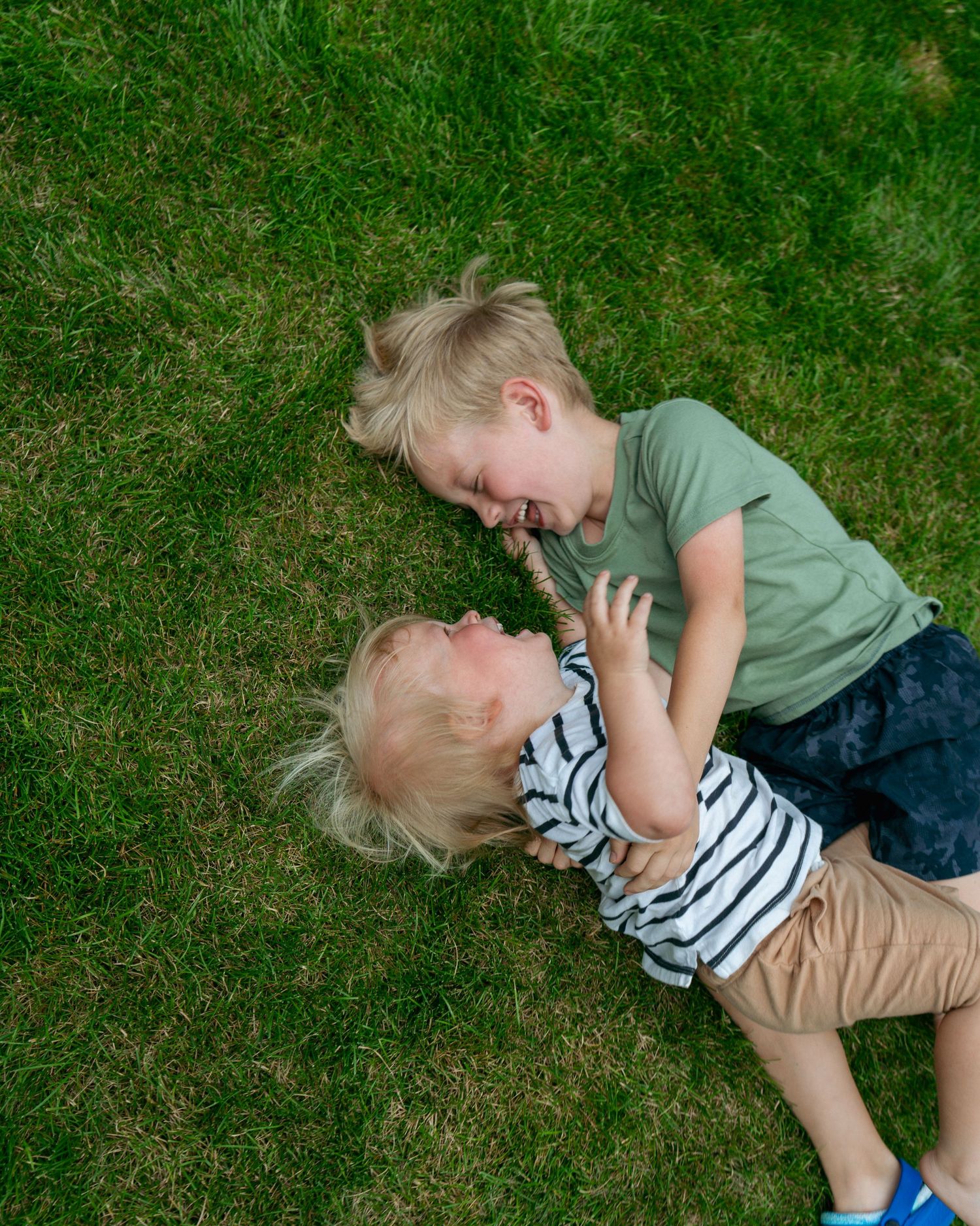 Two young blonde children laughing and playing together on green grass outdoors.