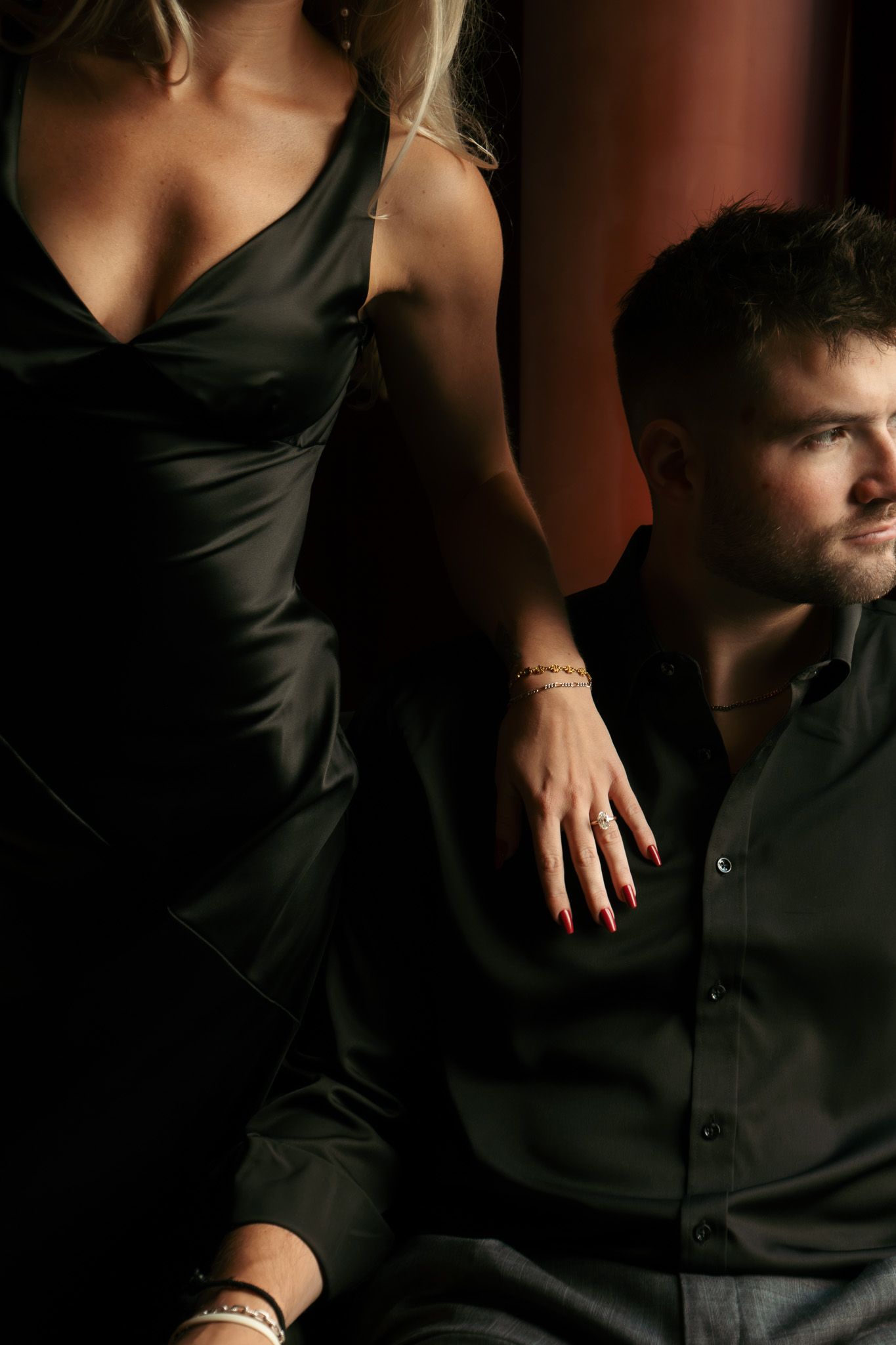A man in a black shirt sits beside a woman in a black satin dress, both exuding elegance in dramatic lighting.