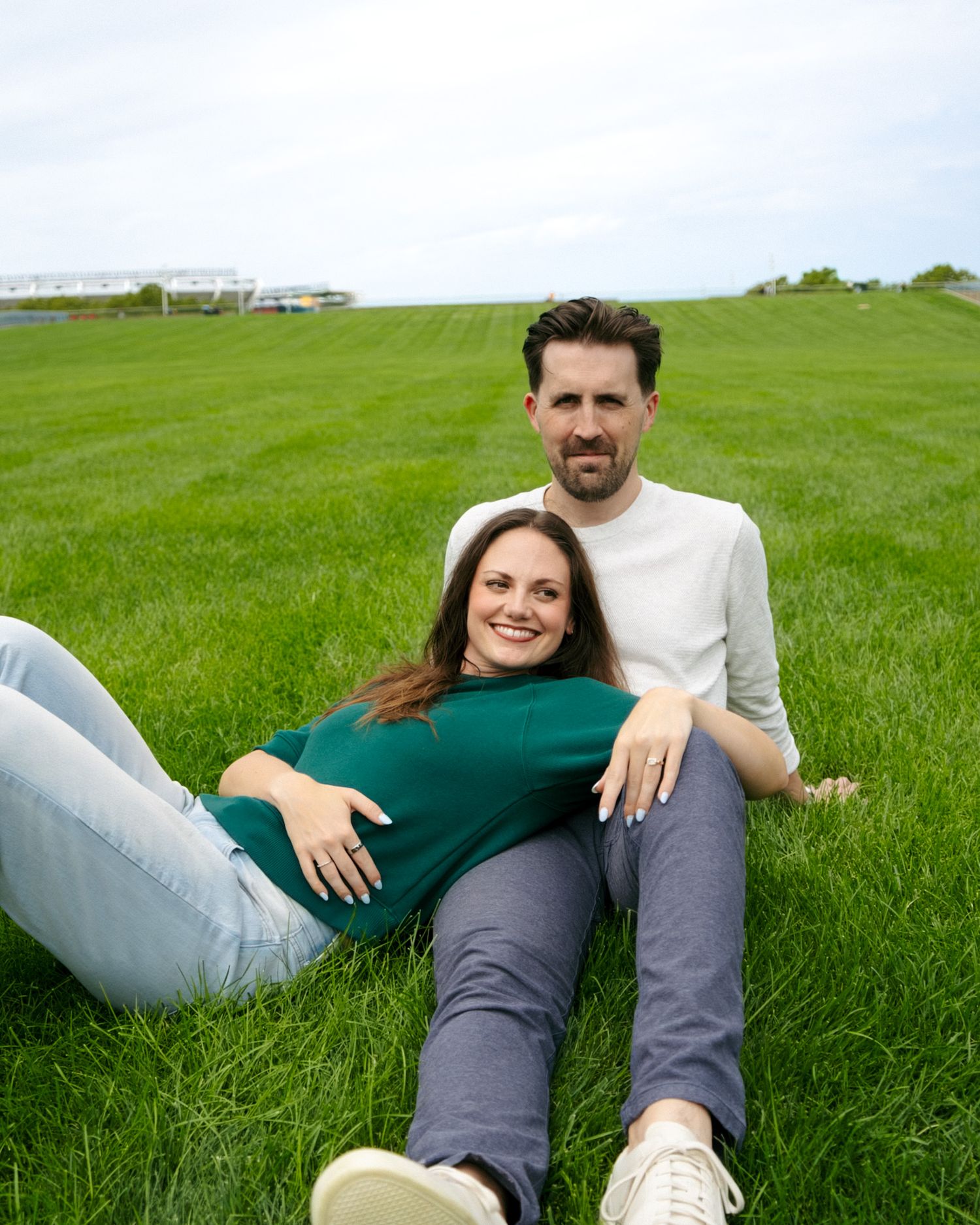 A couple sits together on lush green grass, the woman in green leaning back against the man in a white sweater.