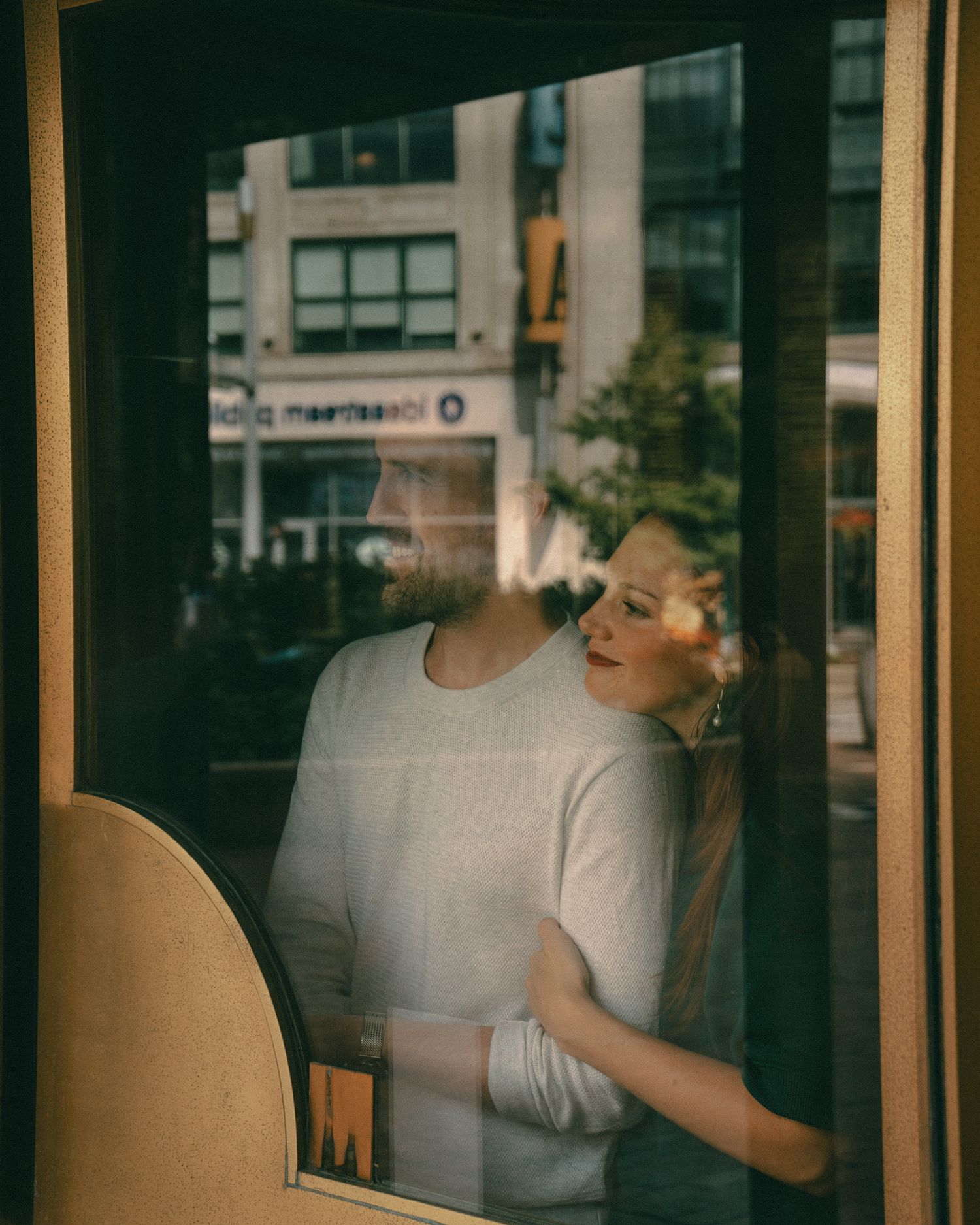 A woman in a white sweater gazes upward, seen through a reflective cafe window on a city street.