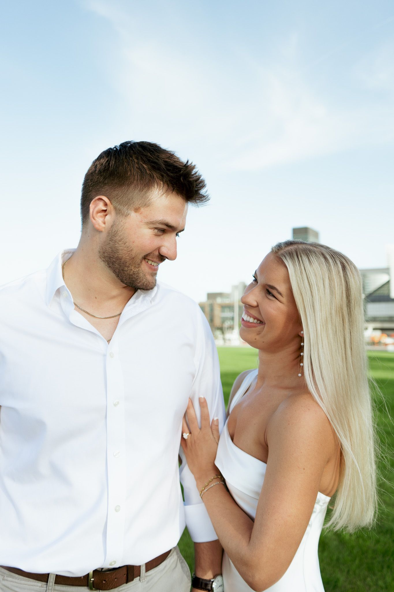 Happy couple in white outfits smiling at each other outdoors on a sunny day with blue sky background.