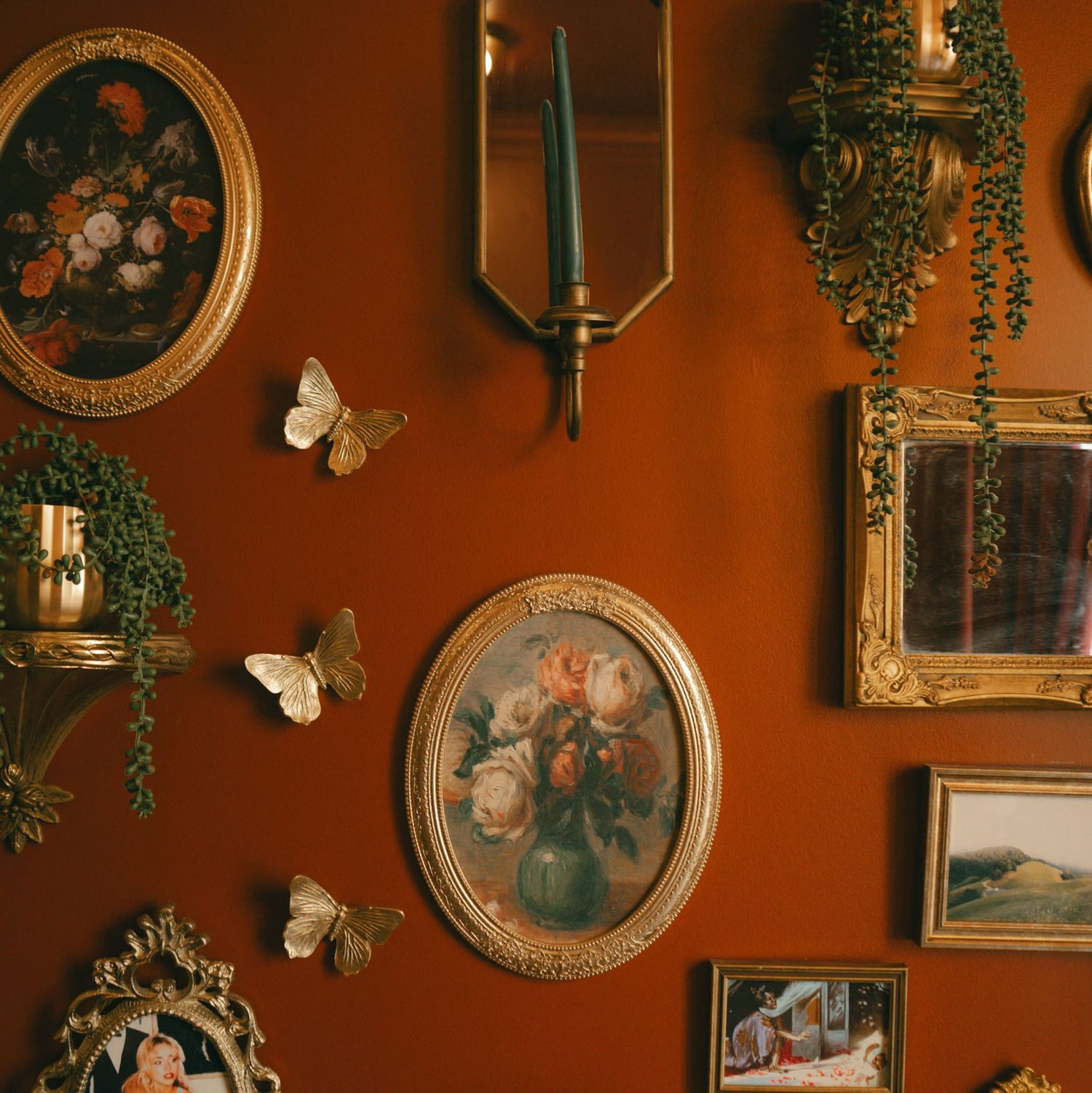 Ornate gold-framed oval paintings and decorative butterflies adorn a rich terracotta wall with candle sconces and trailing greenery.