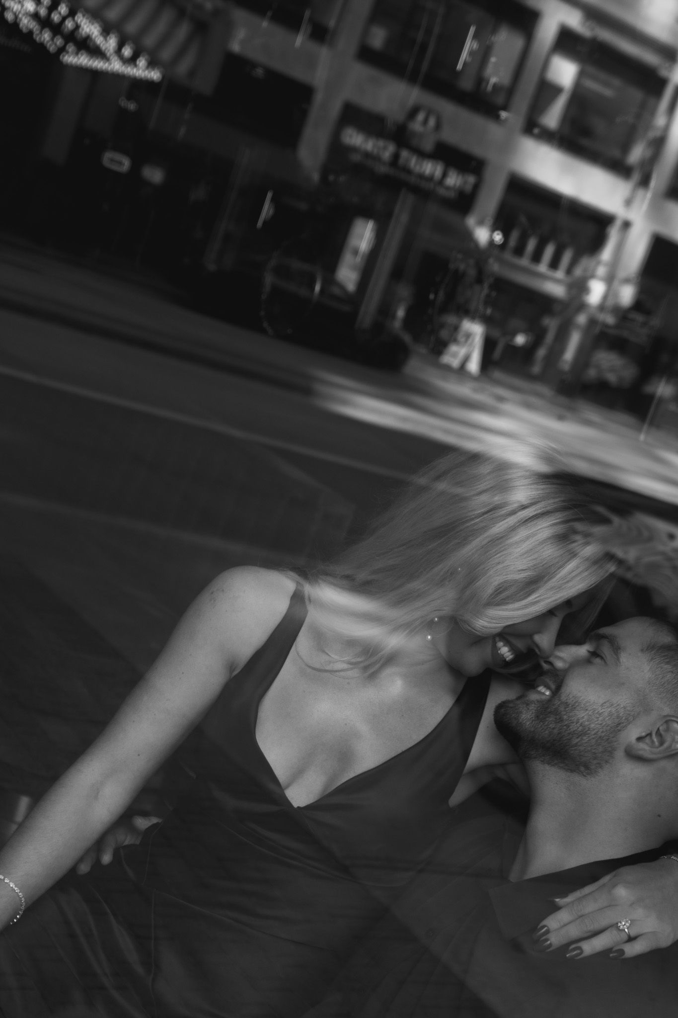 Black and white photo of a couple sharing a romantic kiss on a city street, woman in elegant dress with flowing blonde hair.