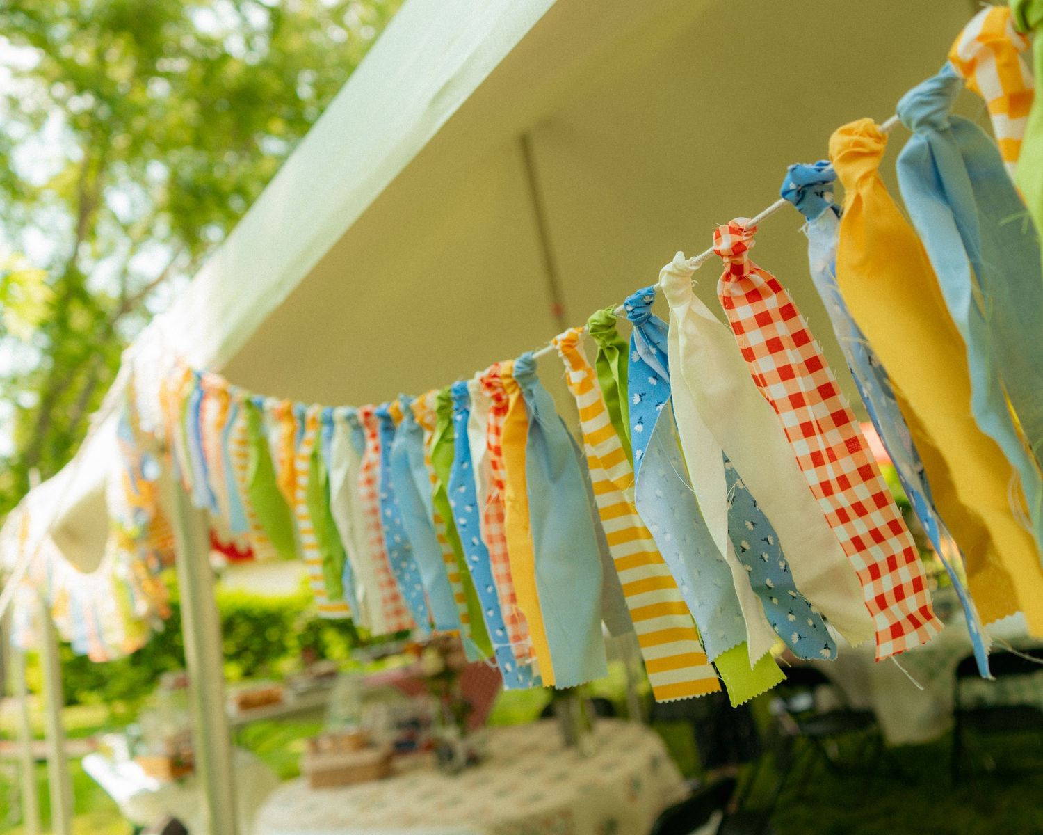 Colorful fabric bunting garland with red, yellow, blue, and green strips hanging under a white tent outdoors.
