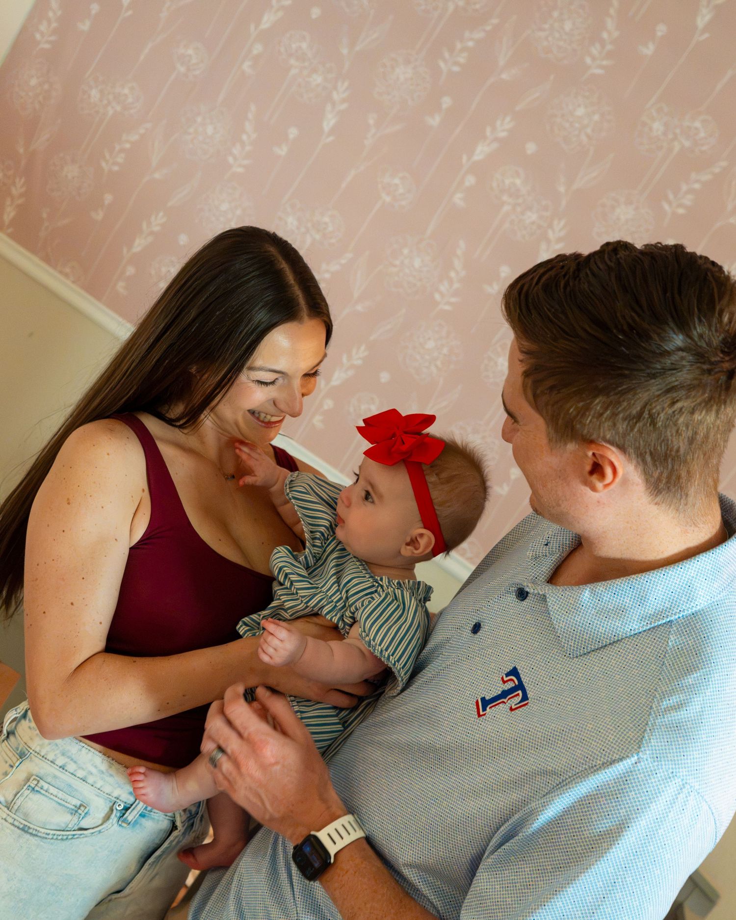 Young couple smiling at their baby girl wearing a red bow headband against a pink floral wallpaper backdrop.