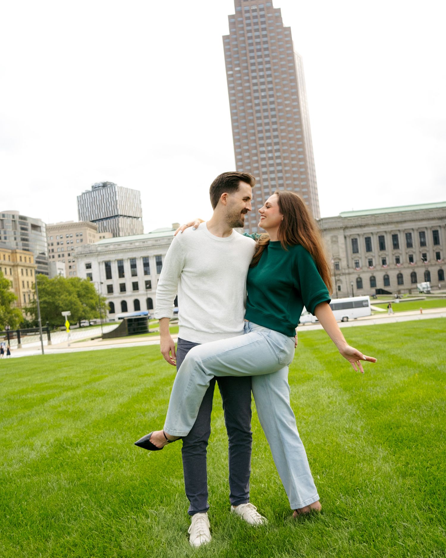 A happy couple laughing and dancing on green grass with city buildings in the background.