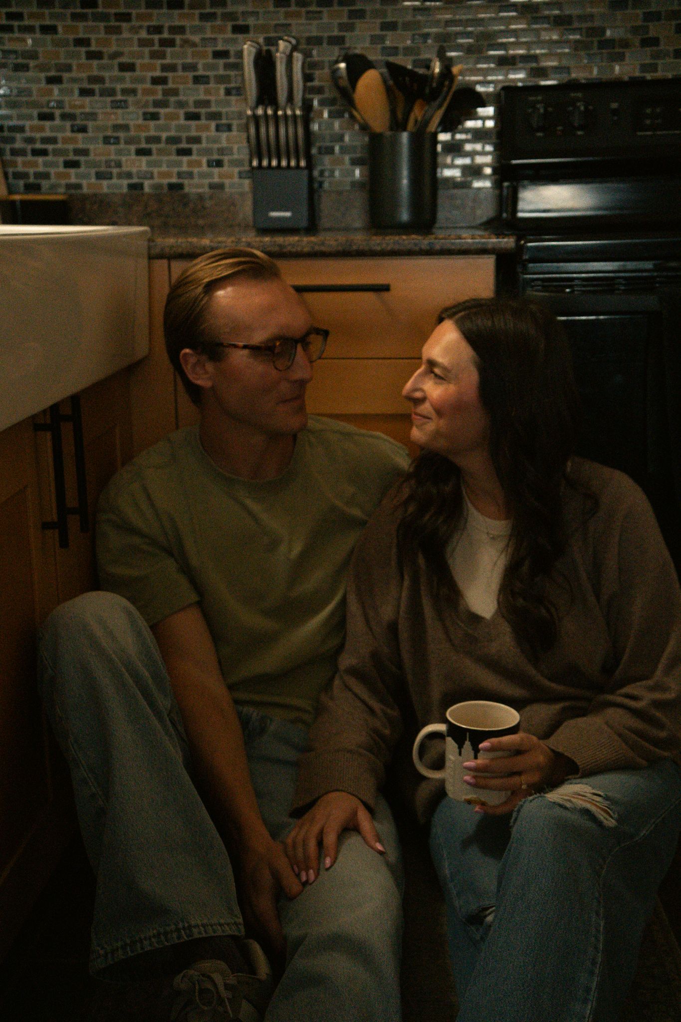 A couple sits on a kitchen floor, smiling at each other, woman holding a mug, man wearing glasses and olive shirt.