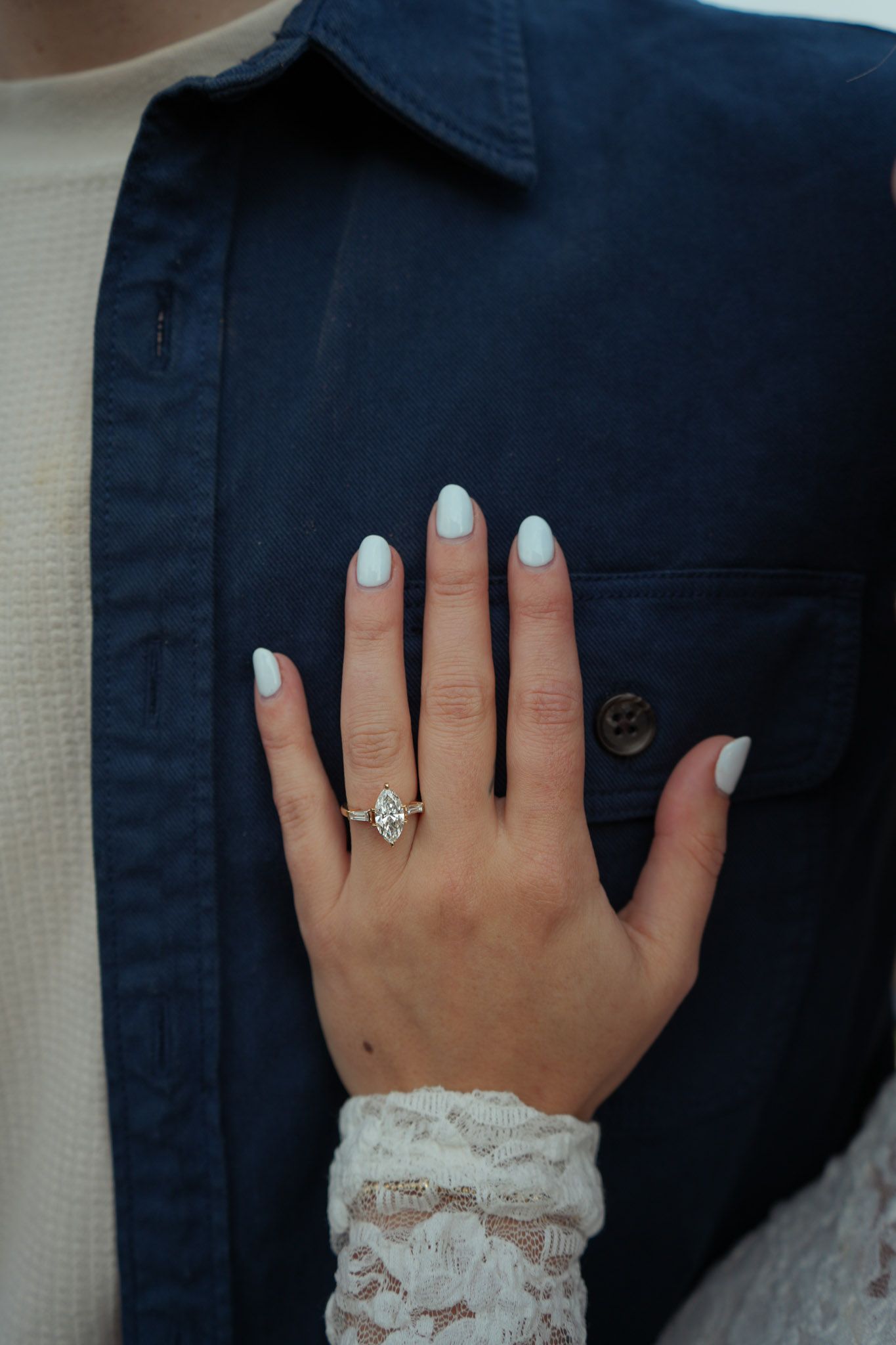 Close-up of a hand with light blue nails displaying a diamond engagement ring, resting on a navy blue shirt.