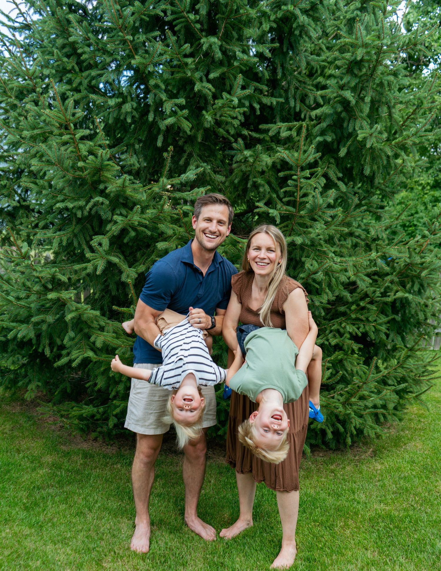 A smiling couple holds two young children upside down in front of a lush green evergreen tree outdoors.