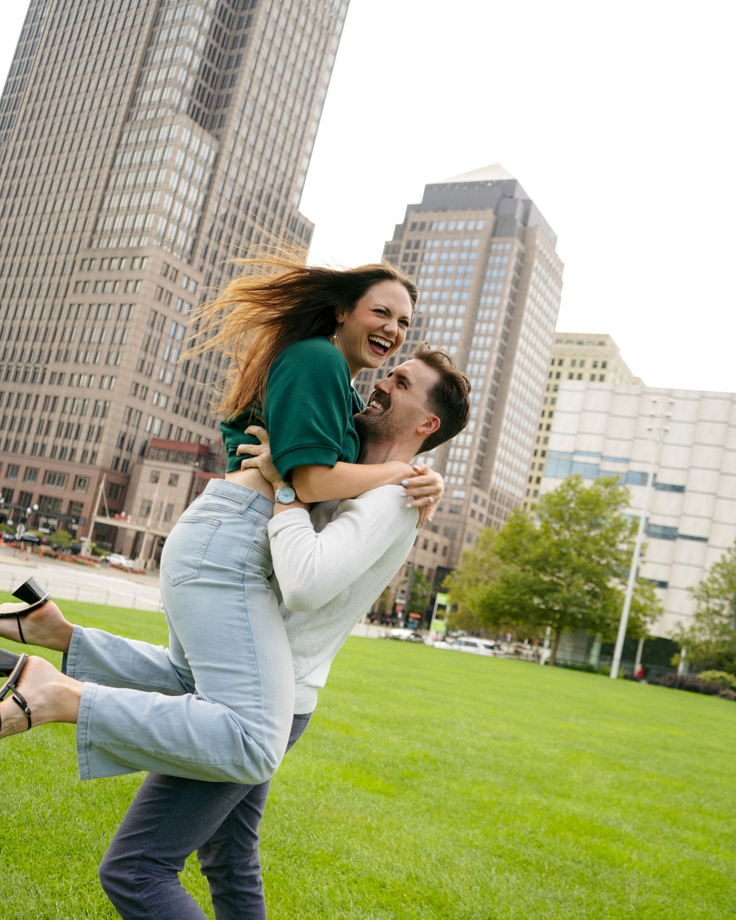 A joyful couple laughing as man lifts woman in his arms on green lawn with city skyscrapers behind.