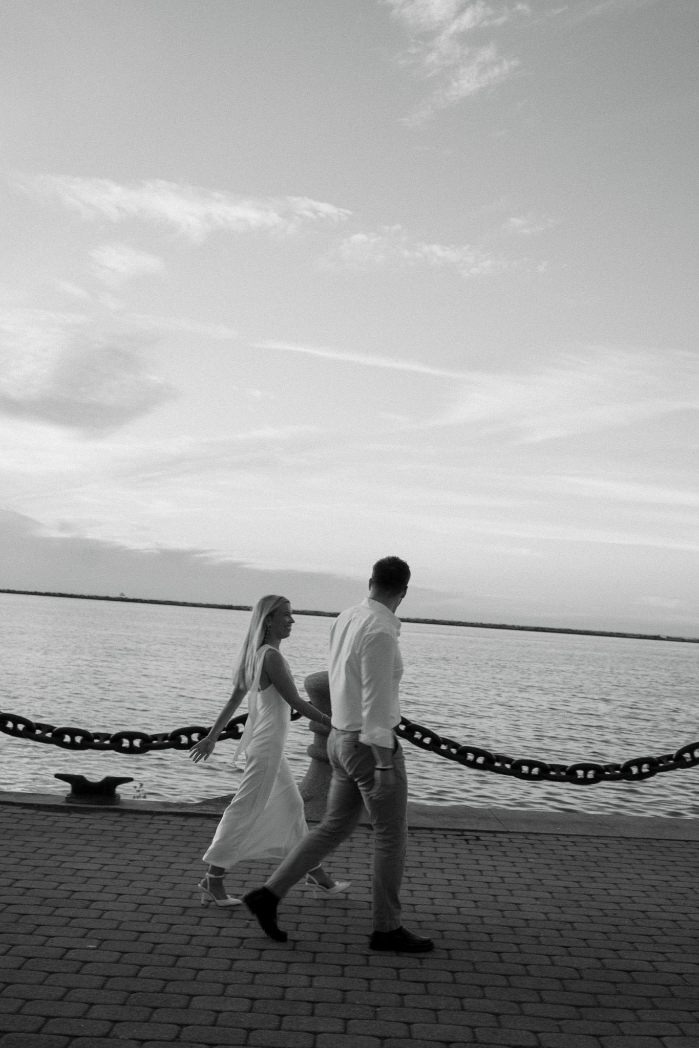 Black and white photo of a bride and groom walking hand in hand along a waterfront promenade at sunset.