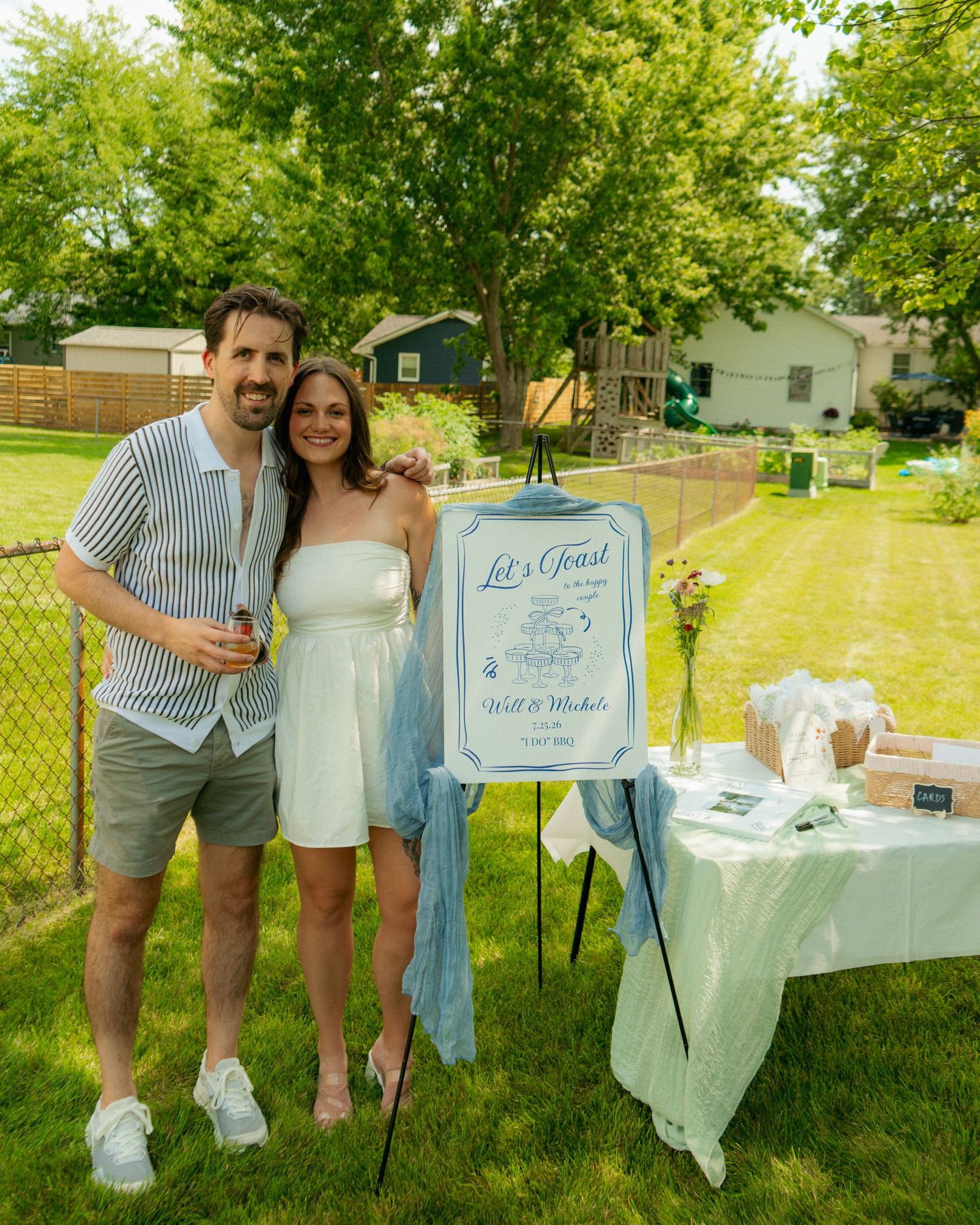 Couple posing beside a Let's Toast celebration sign on an easel at an outdoor backyard party.
