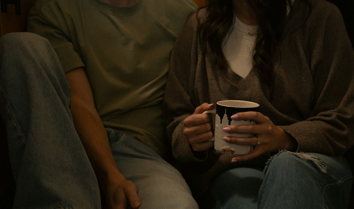 Two people sitting together on a couch, sharing a warm mug, in a cozy dimly lit setting.