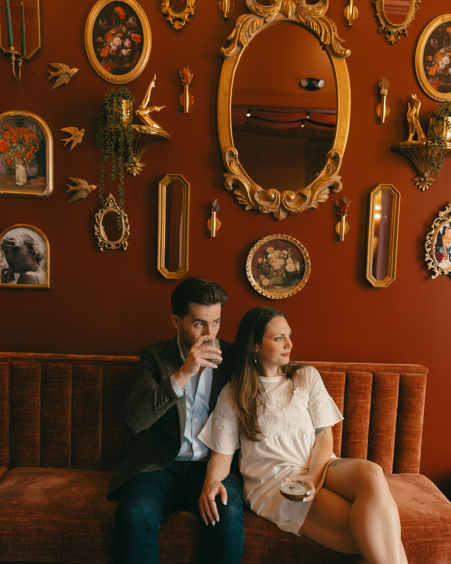 Couple sitting on velvet sofa in moody bar with ornate gold mirrors and vintage paintings on deep red wall.