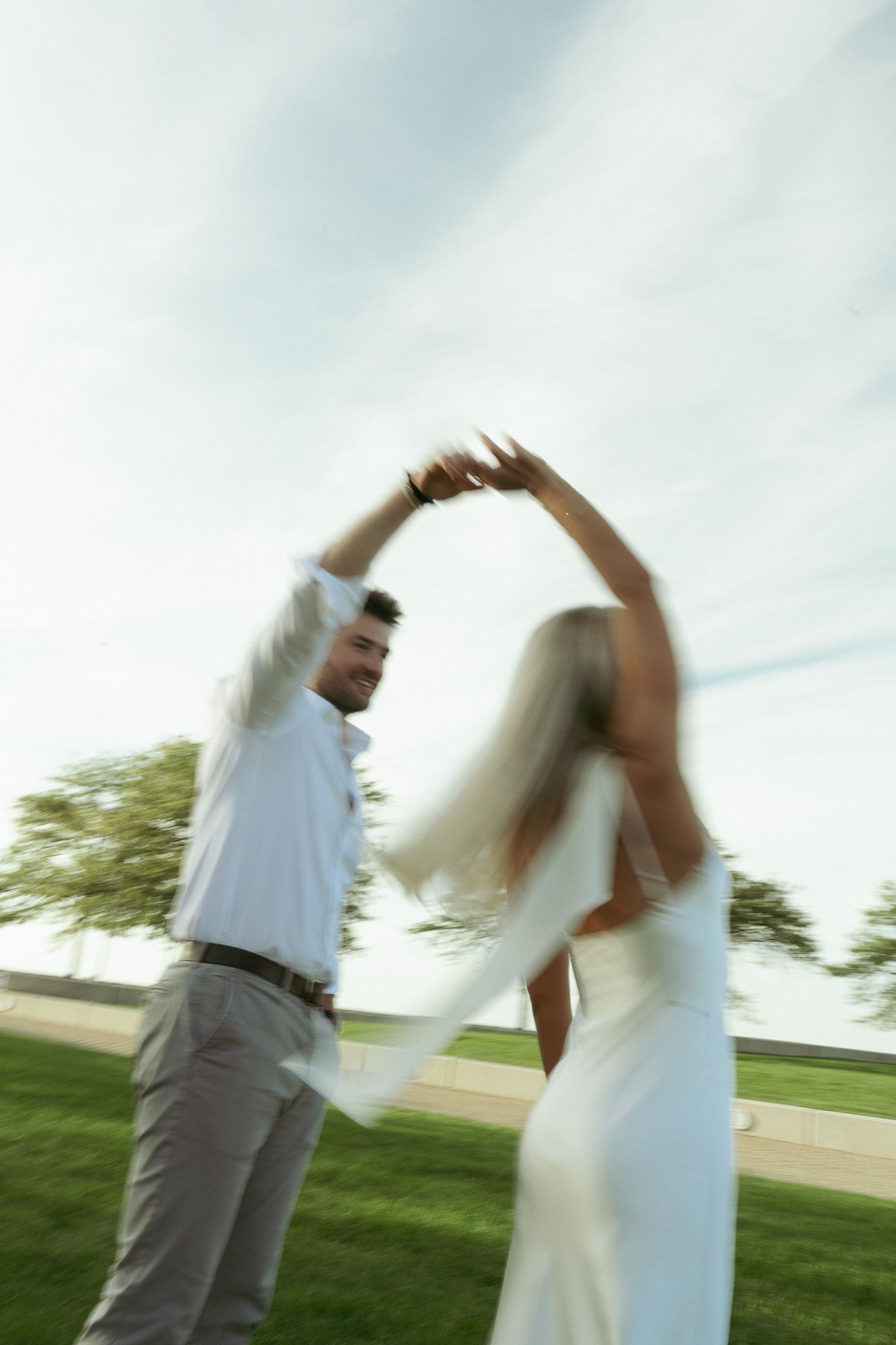 A couple in wedding attire dances outdoors on green grass, captured with motion blur under a cloudy sky.