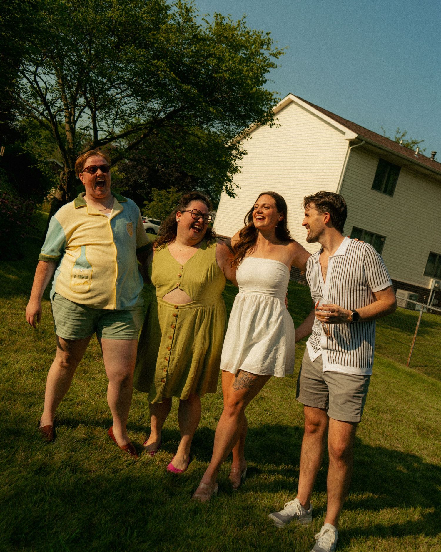 Four friends laughing and walking together on a sunny green lawn beside a white house on a warm summer day.