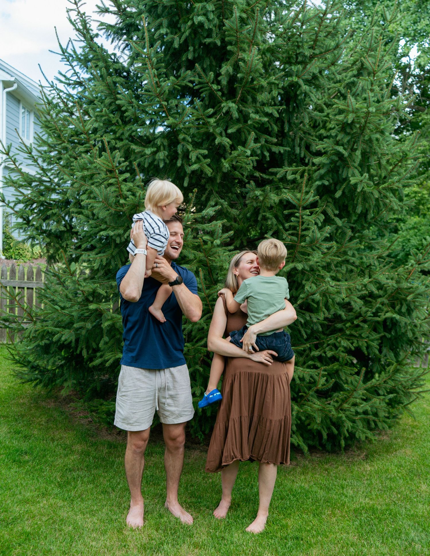 A happy family of four poses barefoot on green grass in front of tall evergreen trees on a sunny day.