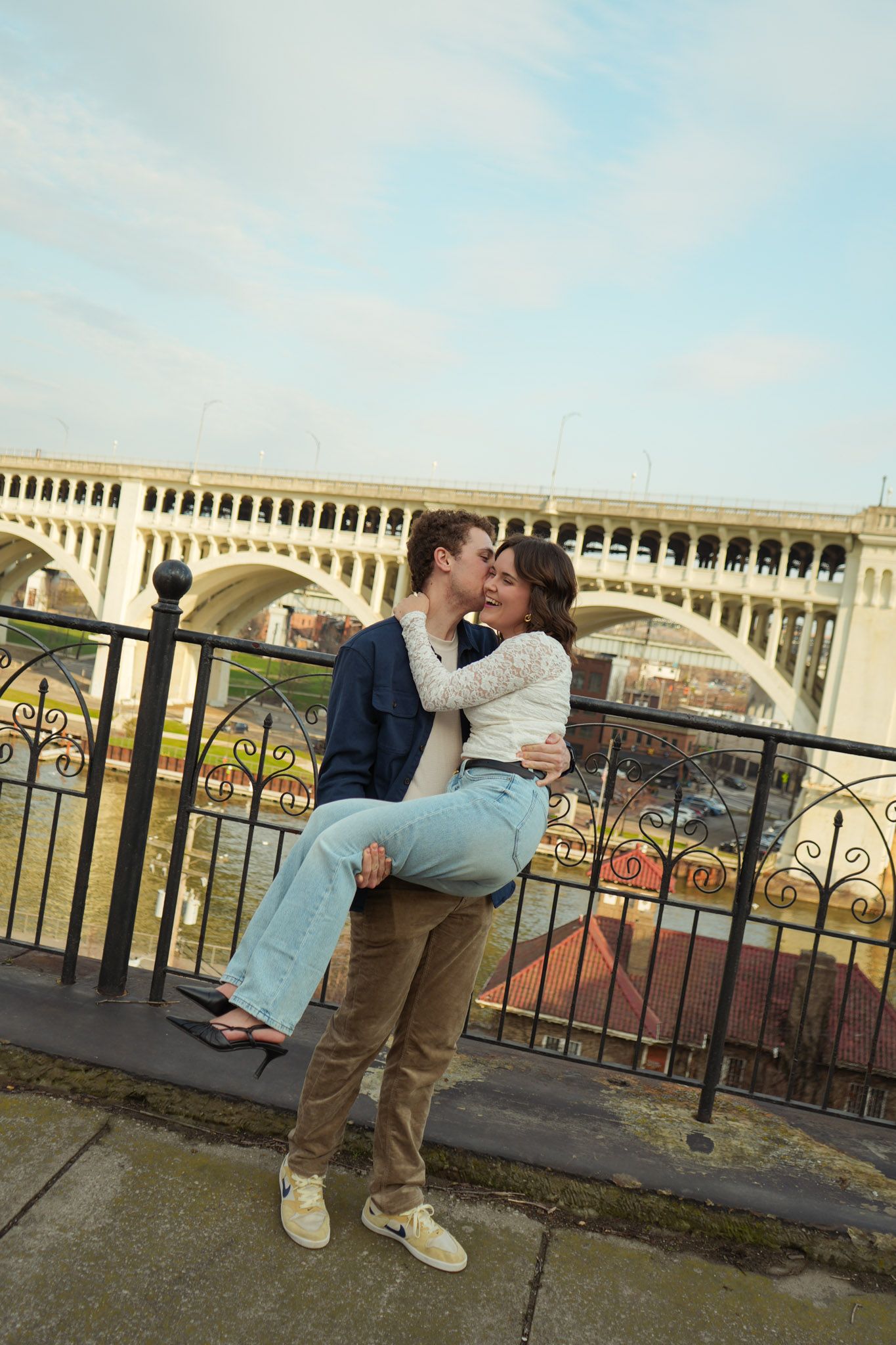 A couple shares a romantic kiss against an ornate bridge railing overlooking a scenic cityscape on an overcast day.