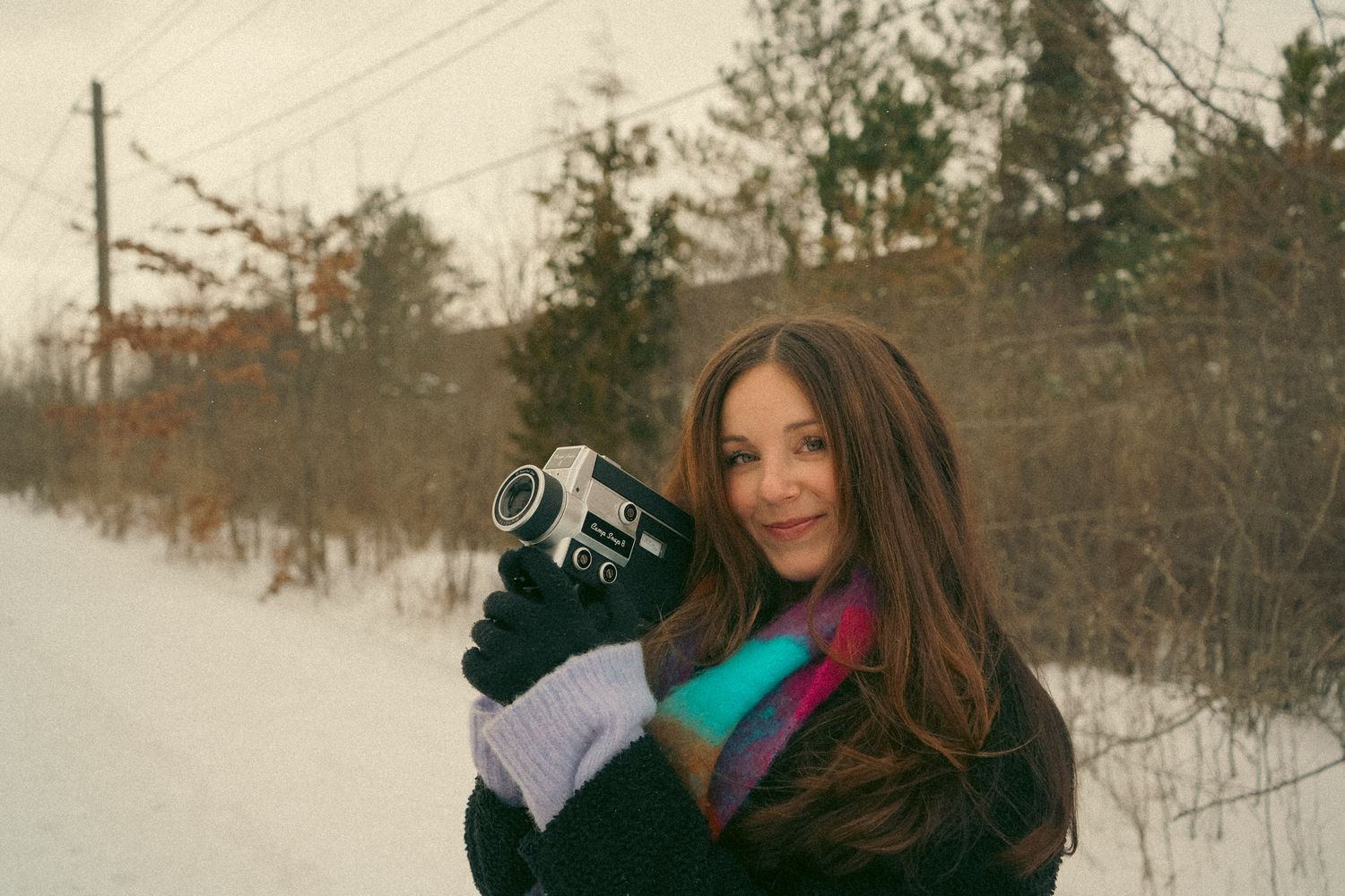 A person wearing a winter hat and scarf stands in a snowy outdoor setting with trees in the background.