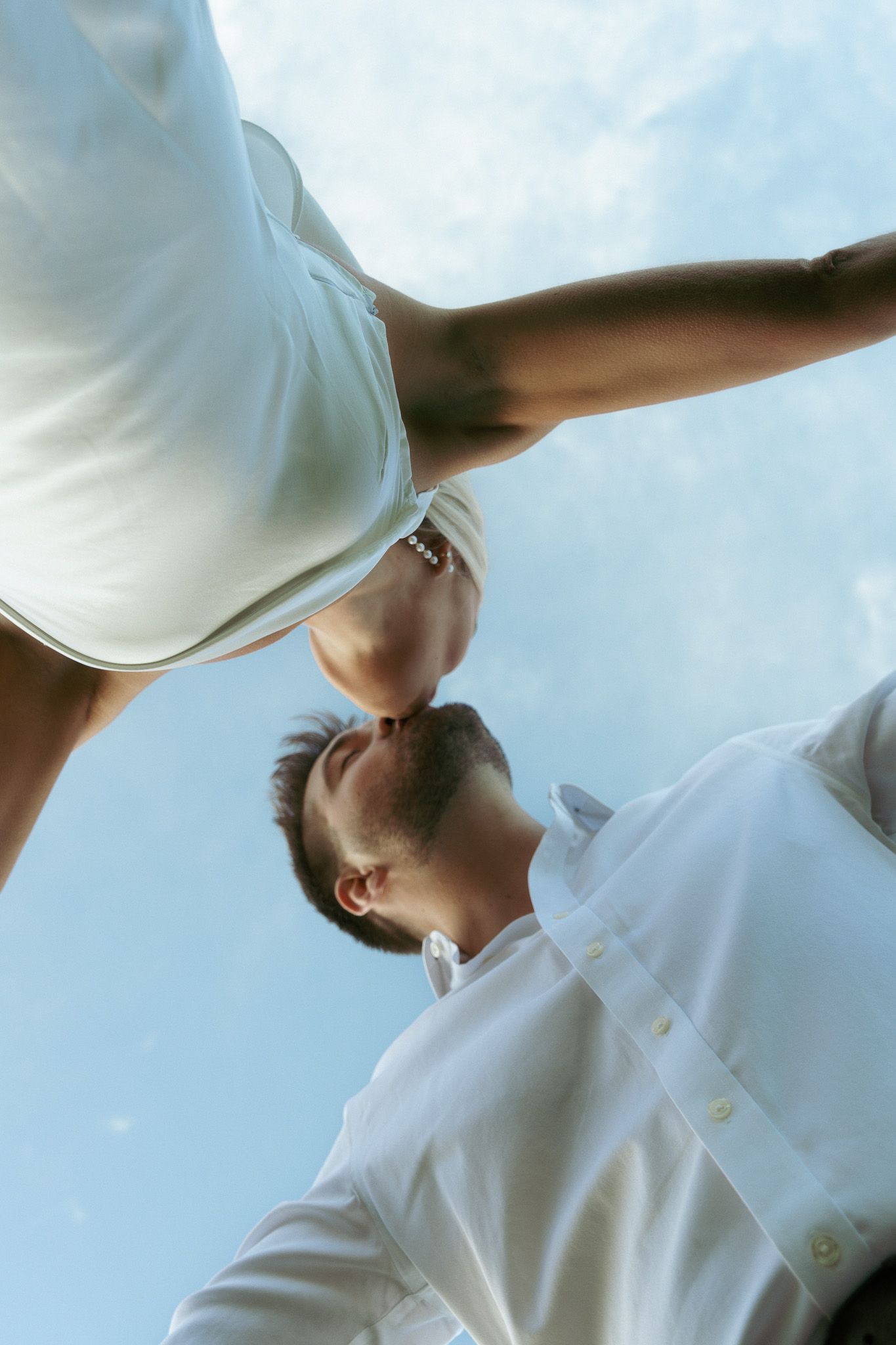 Low angle view of a couple in white outfits sharing a kiss against a blue sky backdrop.