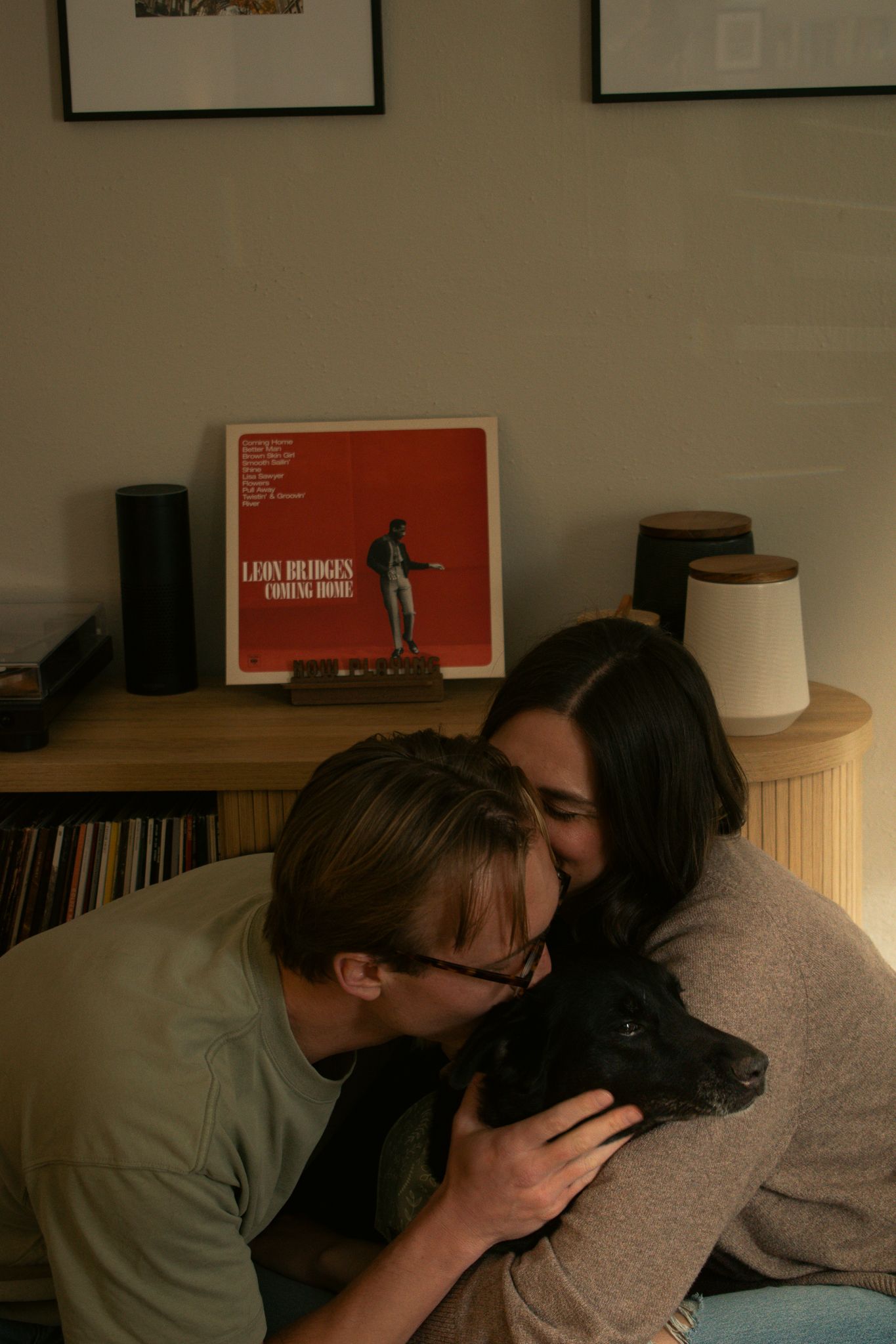 Couple kissing a black dog near a Leon Bridges Coming Home vinyl record on a wooden shelf with speakers.