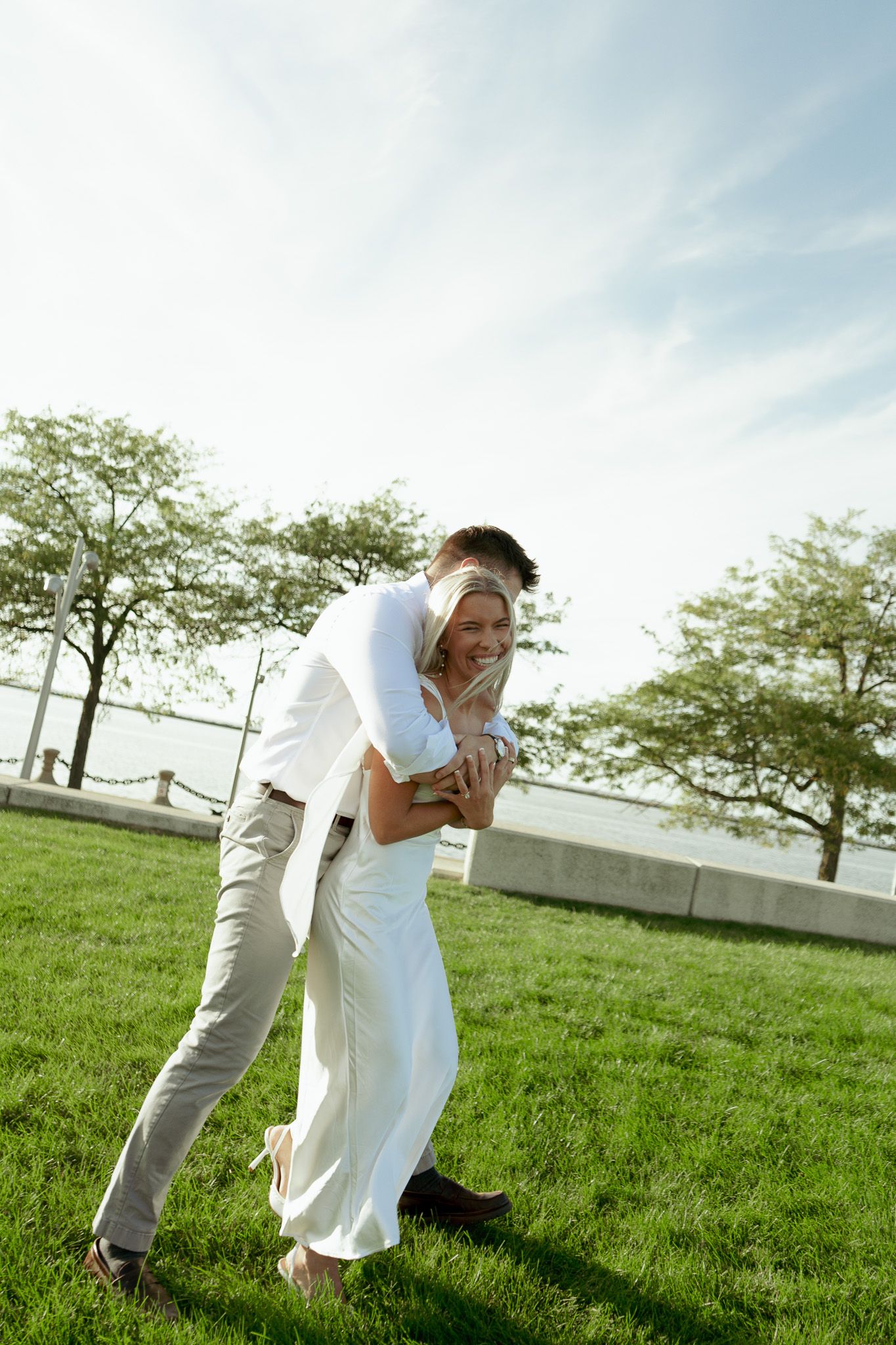 A joyful couple in white wedding attire laughing and embracing on a green lawn with trees and water in the background.