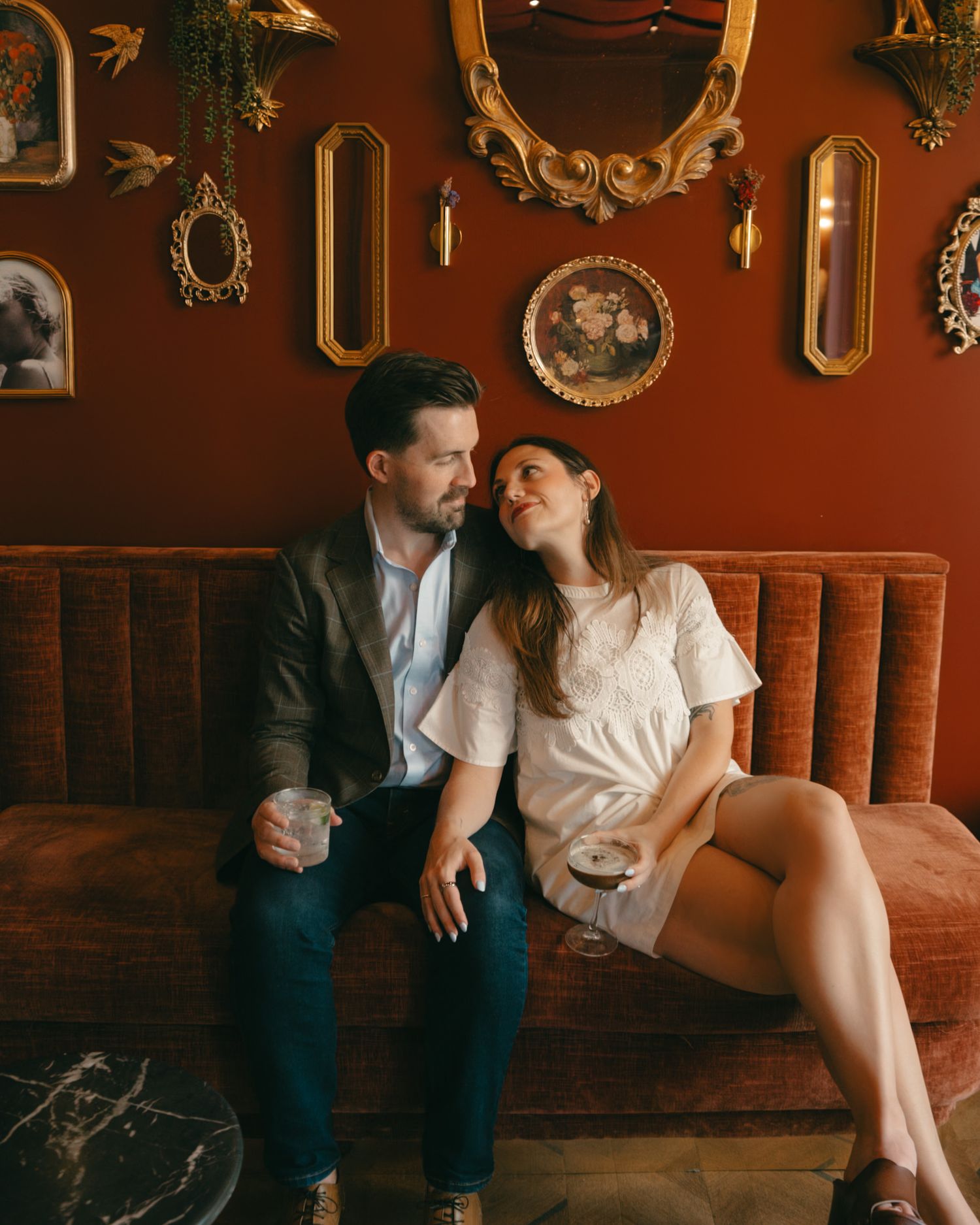 Couple sharing drinks on a velvet sofa, surrounded by ornate gold mirrors on a rich red wall.