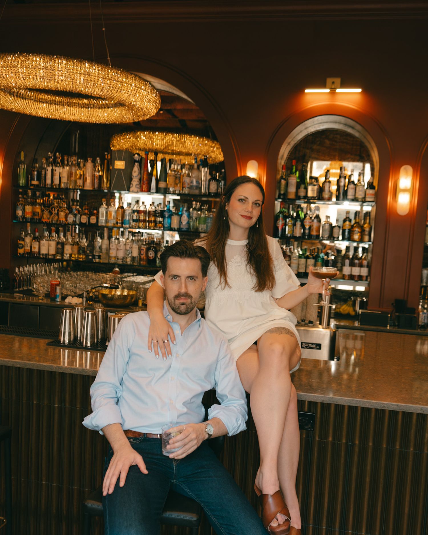 Couple posing at an upscale bar with bottle-lined shelves and a gold chandelier overhead.