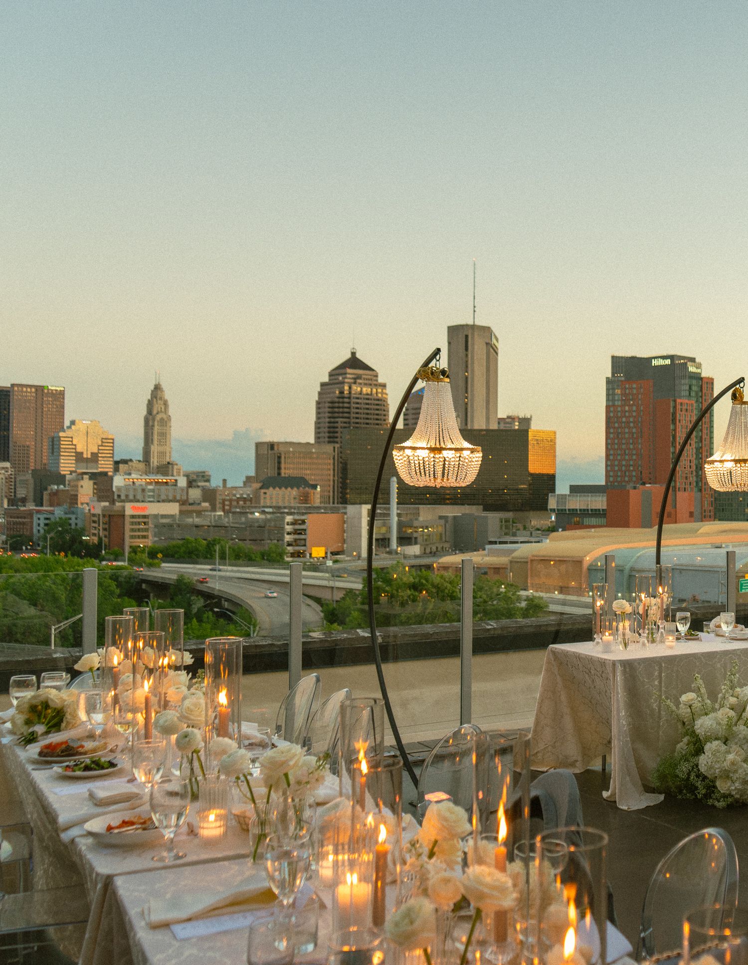 Elegant rooftop wedding reception with chandeliers, candlelit tables, and stunning city skyline backdrop at golden hour.