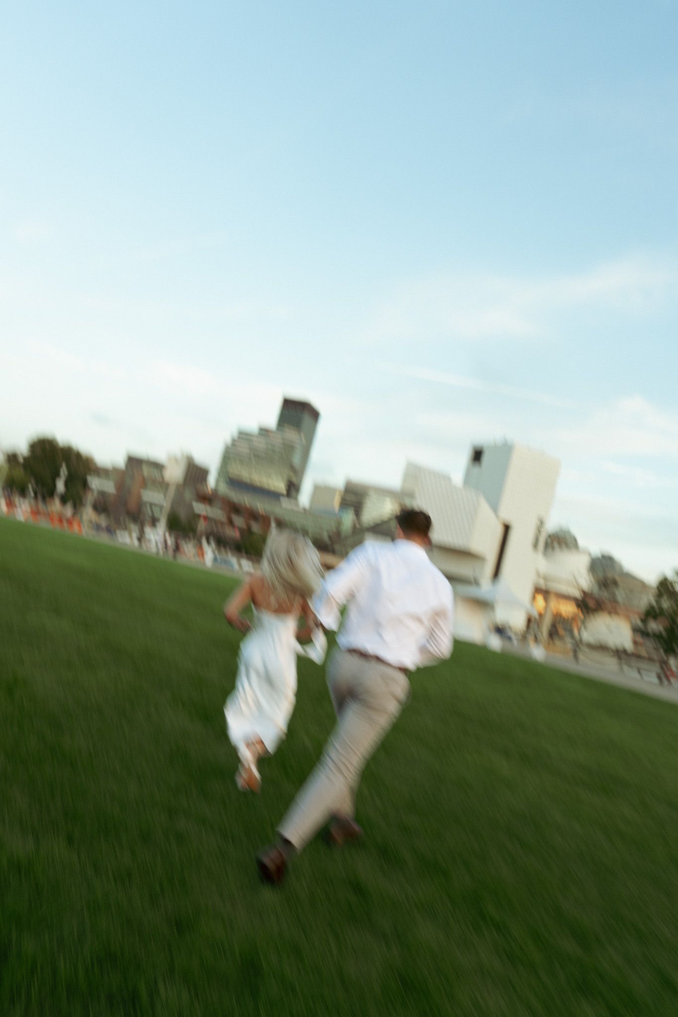 Couple holding hands running on green grass with city skyline in background on a clear day.